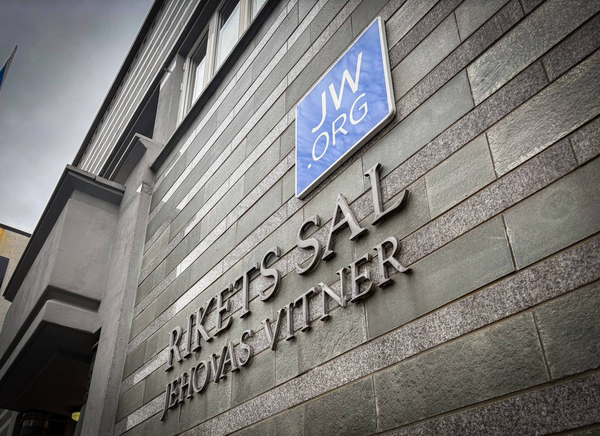 Jehovas Vitner sin møteplass i Kristiansand. Bildet viser en grå murvegg med en blå logo det står JW.org på. Under står det Rikets sal, Jehovas vitner.
