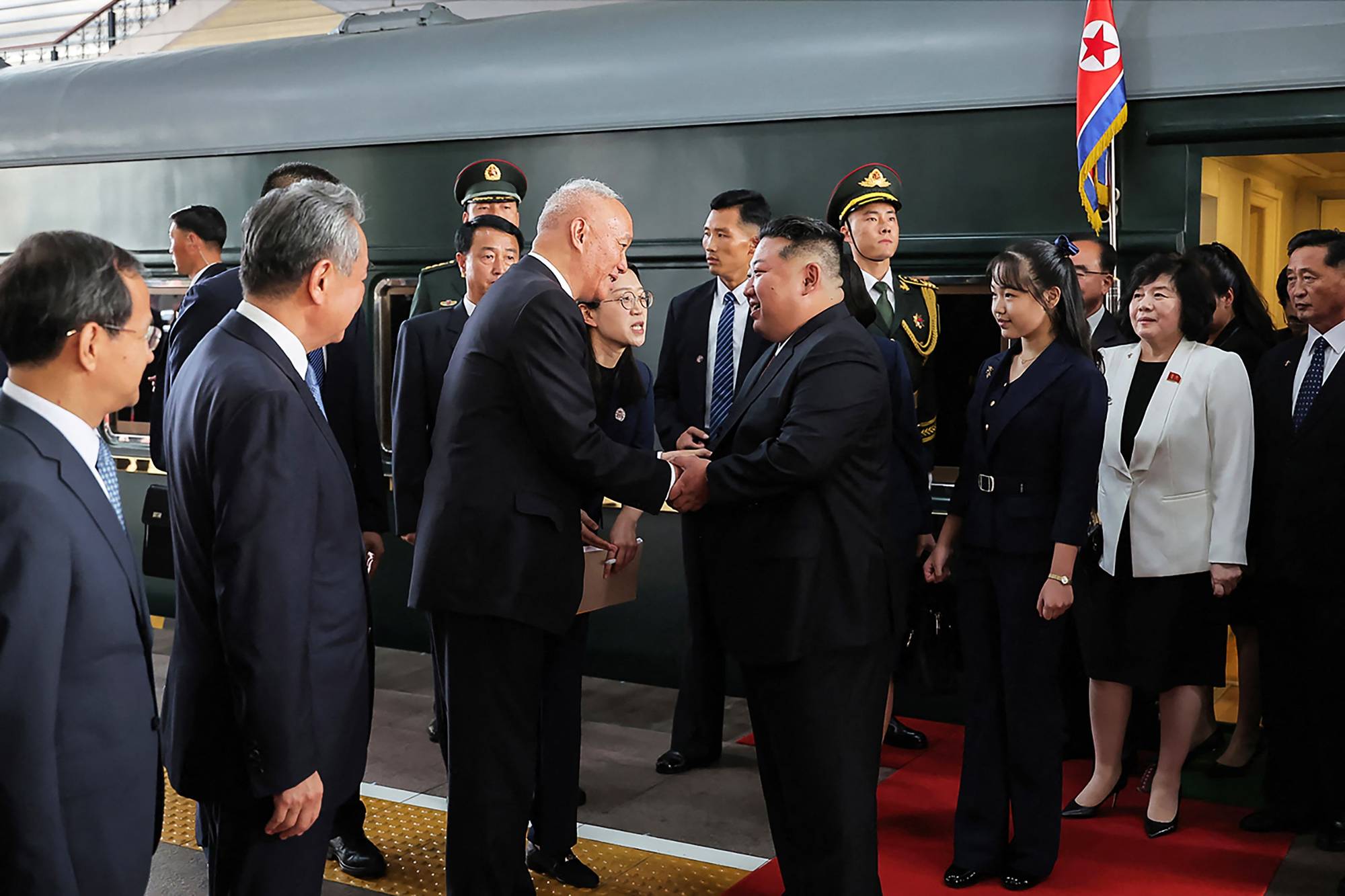 Nord-Koreas leder Kim Jong-un håndhilser på en mann ved et tog. Flere personer er samlet rundt dem, deriblant kvinner og menn i mørke dresser og uniformer. Bakgrunnen viser et tog og en person med nordkoreansk flagg. (Bildebeskrivelsen er laget av en KI-tjeneste)