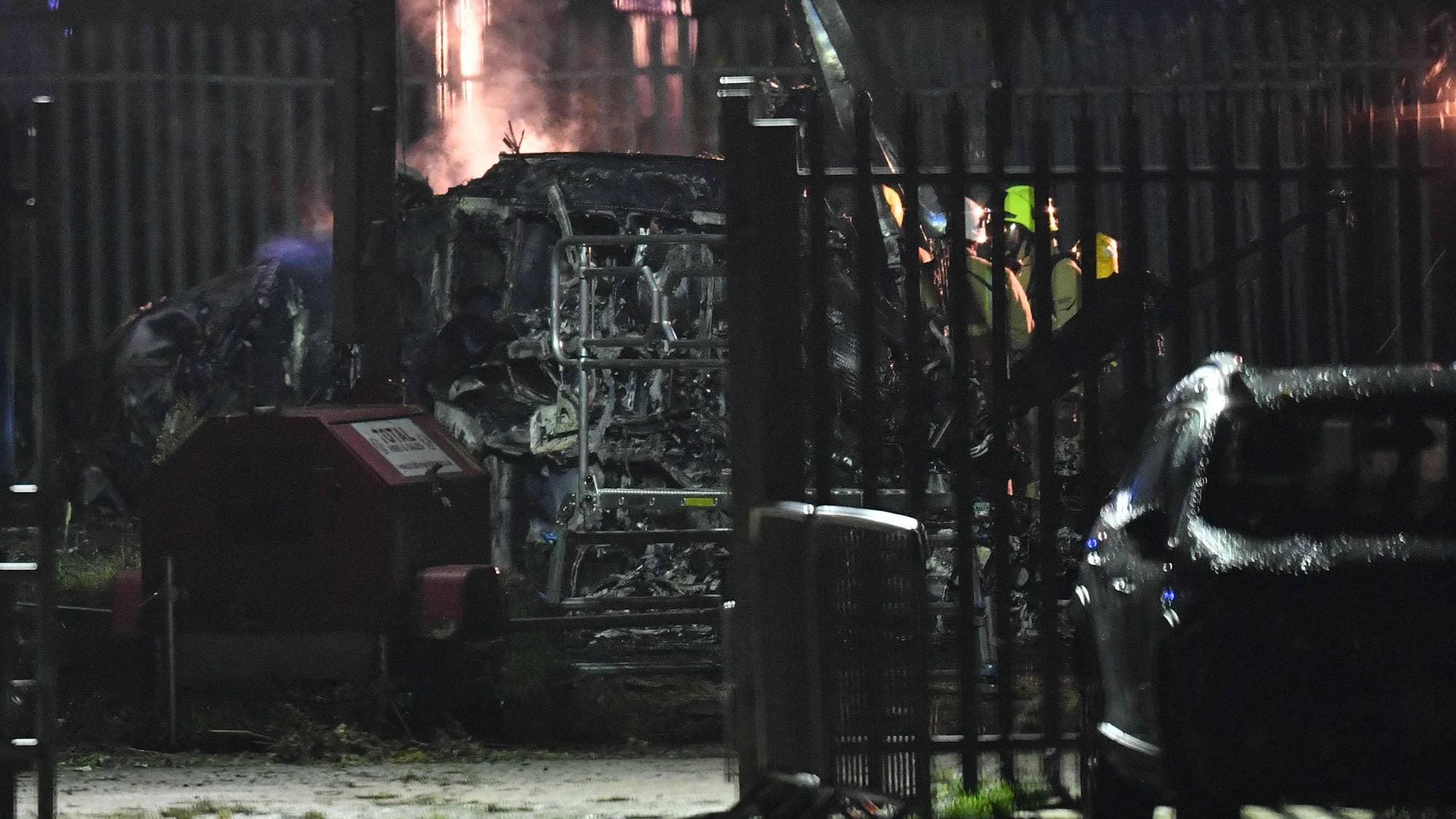 Brannmenn er synlige bak et gjerde, og et brent kjøretøy er synlig. Det er røyk og flammer i bakgrunnen. Bildet er tatt om natten. (Bildebeskrivelsen er laget av en KI-tjeneste)