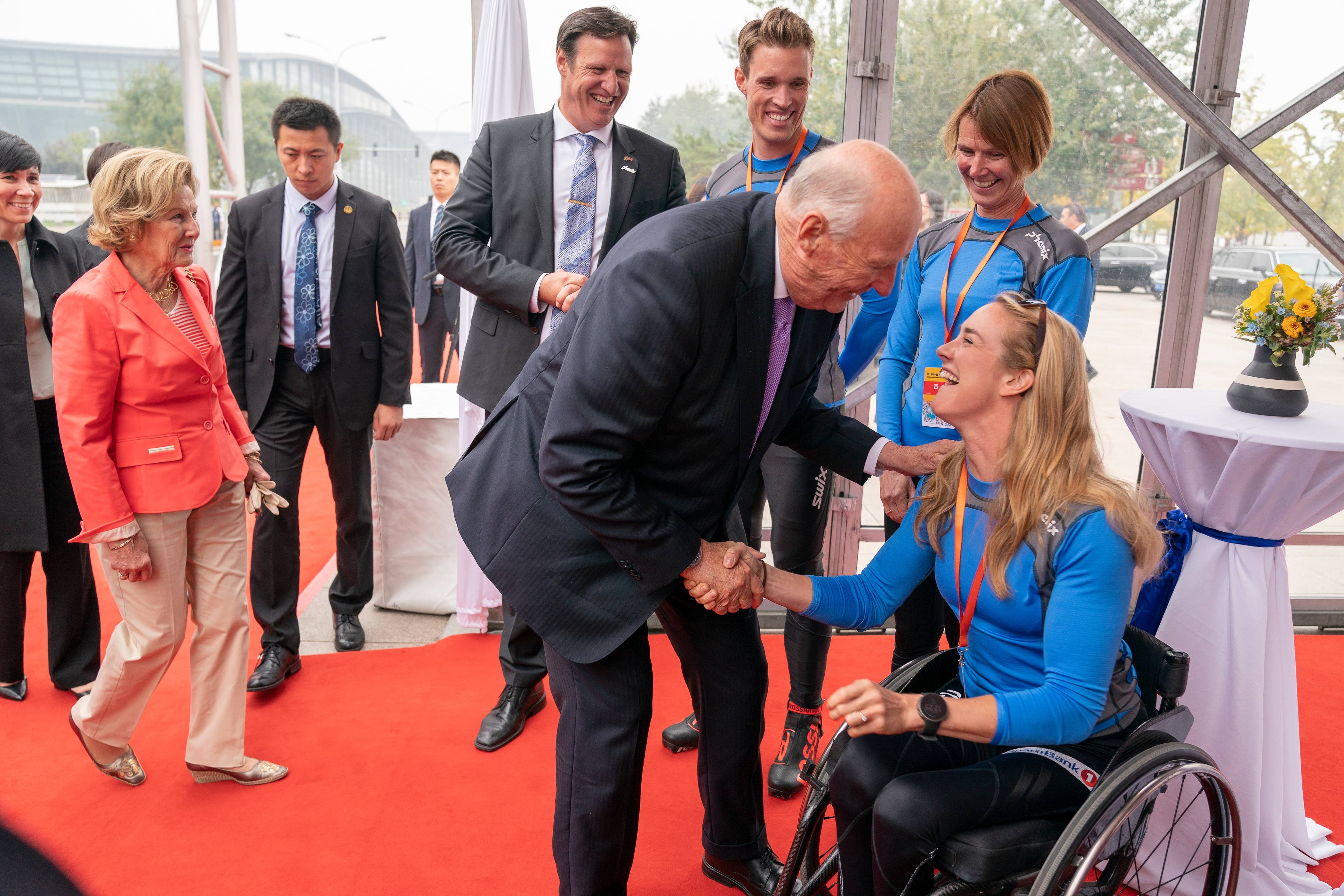 Kong Harald håndhilser på en utøver i rullestol. Dronning Sonja og flere personer står i bakgrunnen. (Bildebeskrivelsen er laget av en KI-tjeneste)