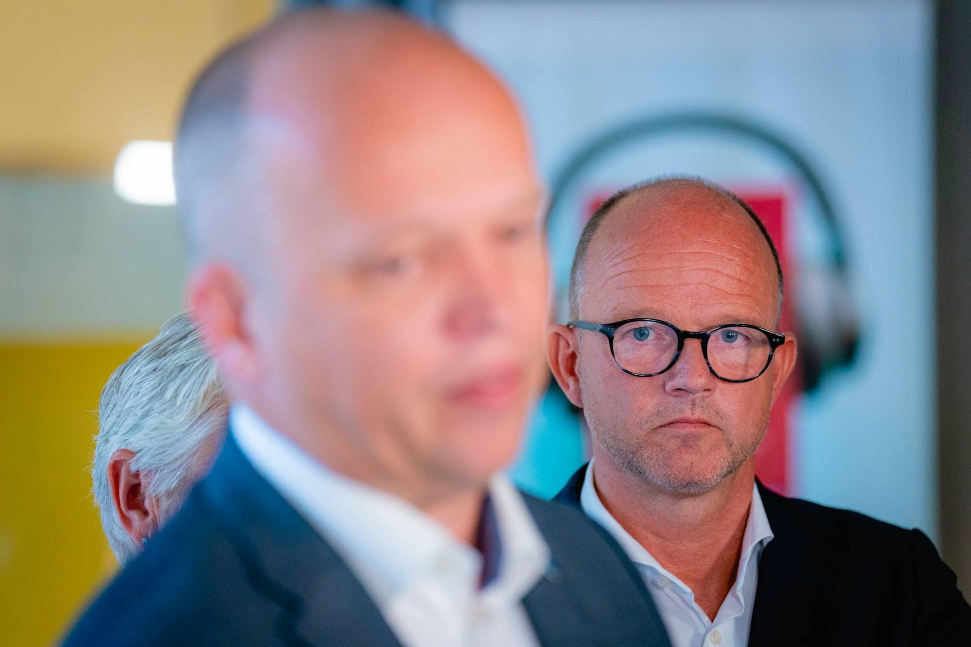 Ole Erik Almlid med briller står bak en person som er ute av fokus. (Bildebeskrivelsen er laget av en KI-tjeneste)