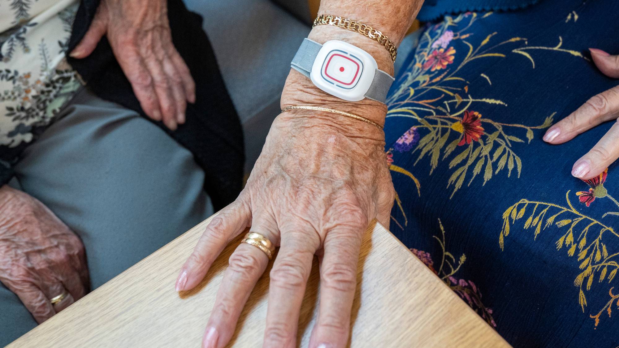 Eldre kvinne med trygghetsalarm på armen sitter ved et bord sammen med en annen person. (Bildebeskrivelsen er laget av en KI-tjeneste)