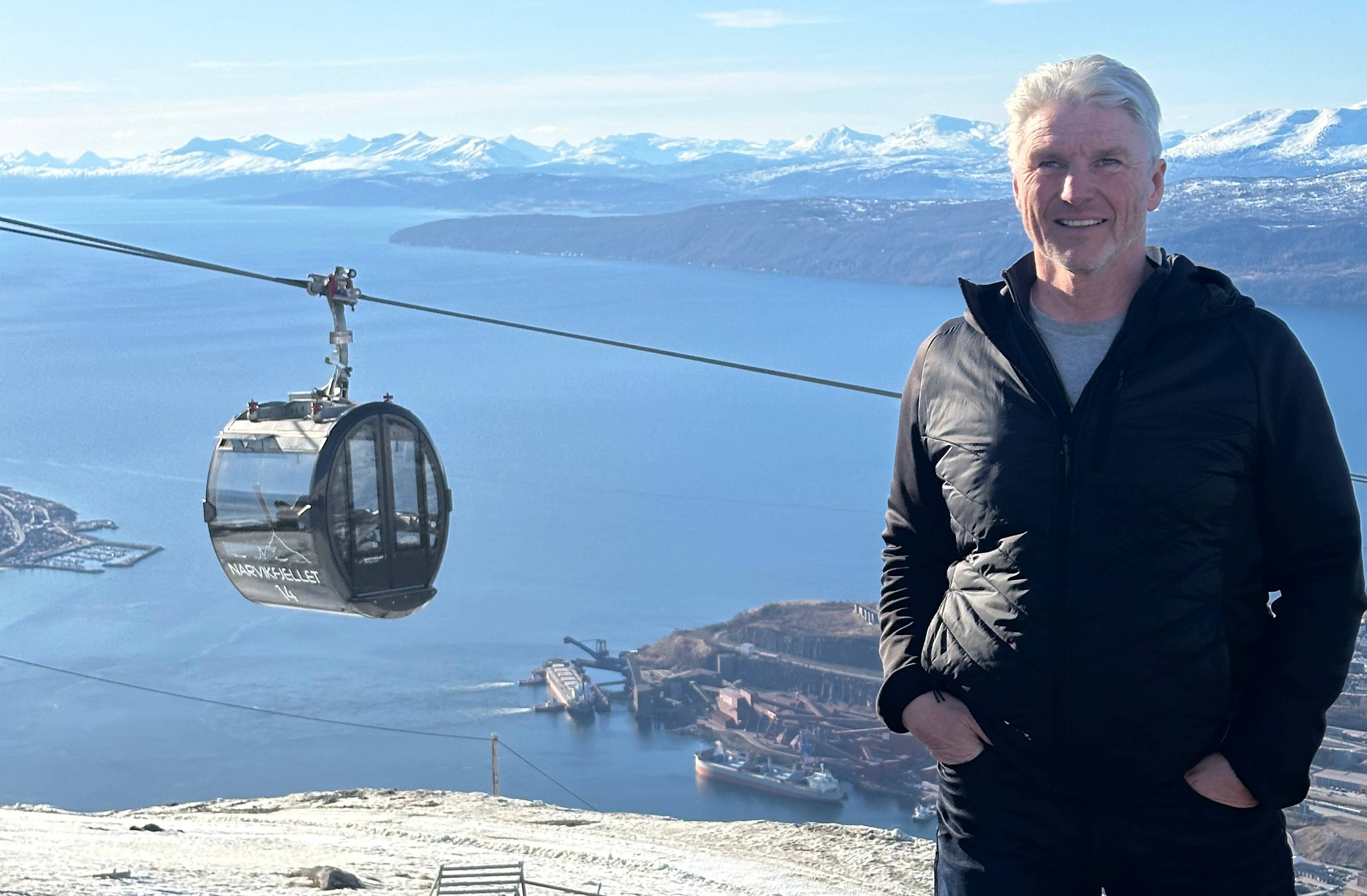 En mann står på en steinete fjellside. I bakgrunnen er det et fjordlandskap med snødekte fjell. En taubane med en gondol henger over fjorden. Ved fjorden ligger det skip og industribygg. (Bildebeskrivelsen er laget av en KI-tjeneste)