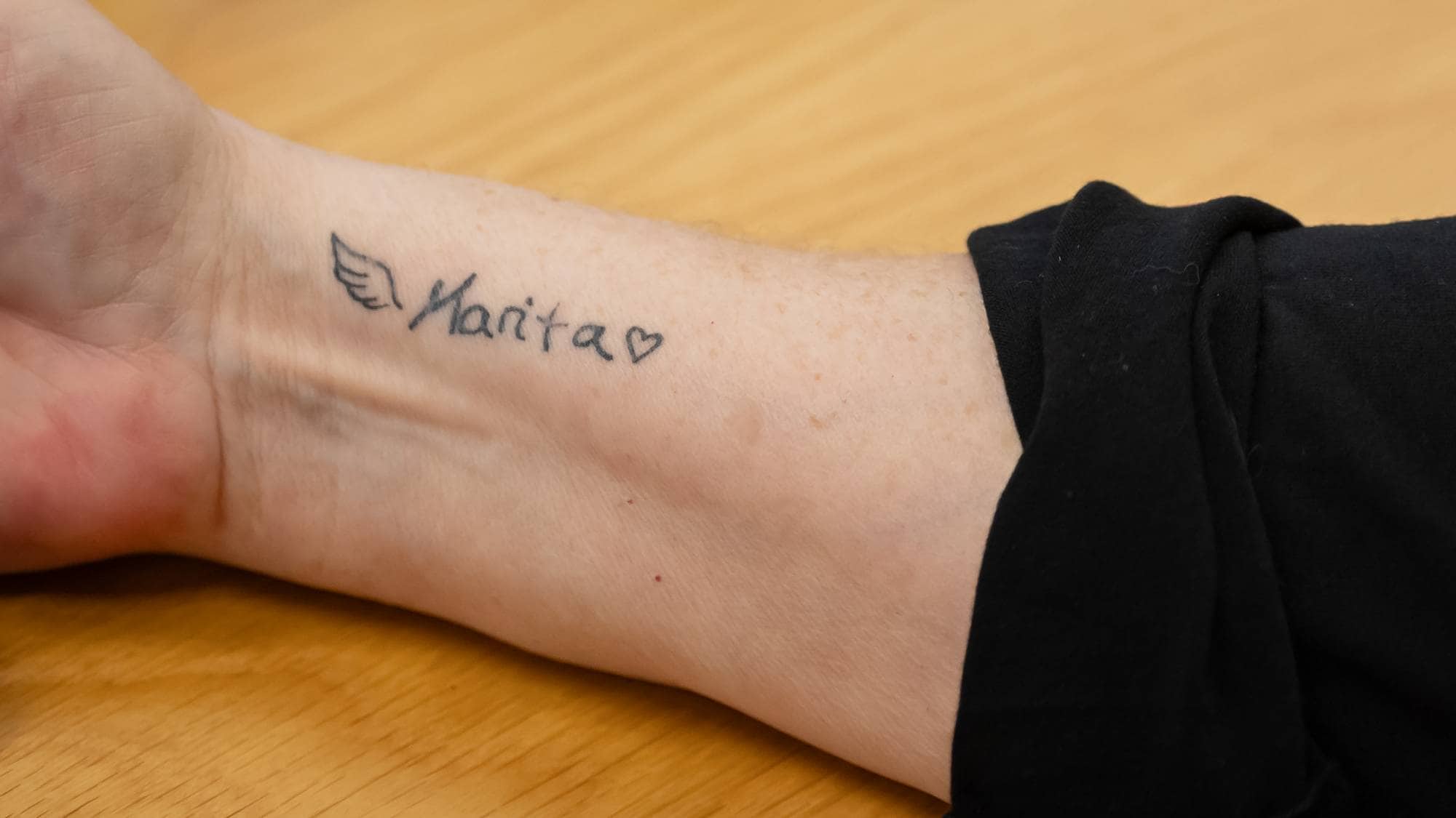 Vi ser kun deler av armen og genseren til en kvinne. Hun har på seg en svart genser. På innsiden av armen har hun tatovert inn navnet på sin datter, "Marita". Til venstre for navnet er et bilde av en englevinge og til høyre et hjerte