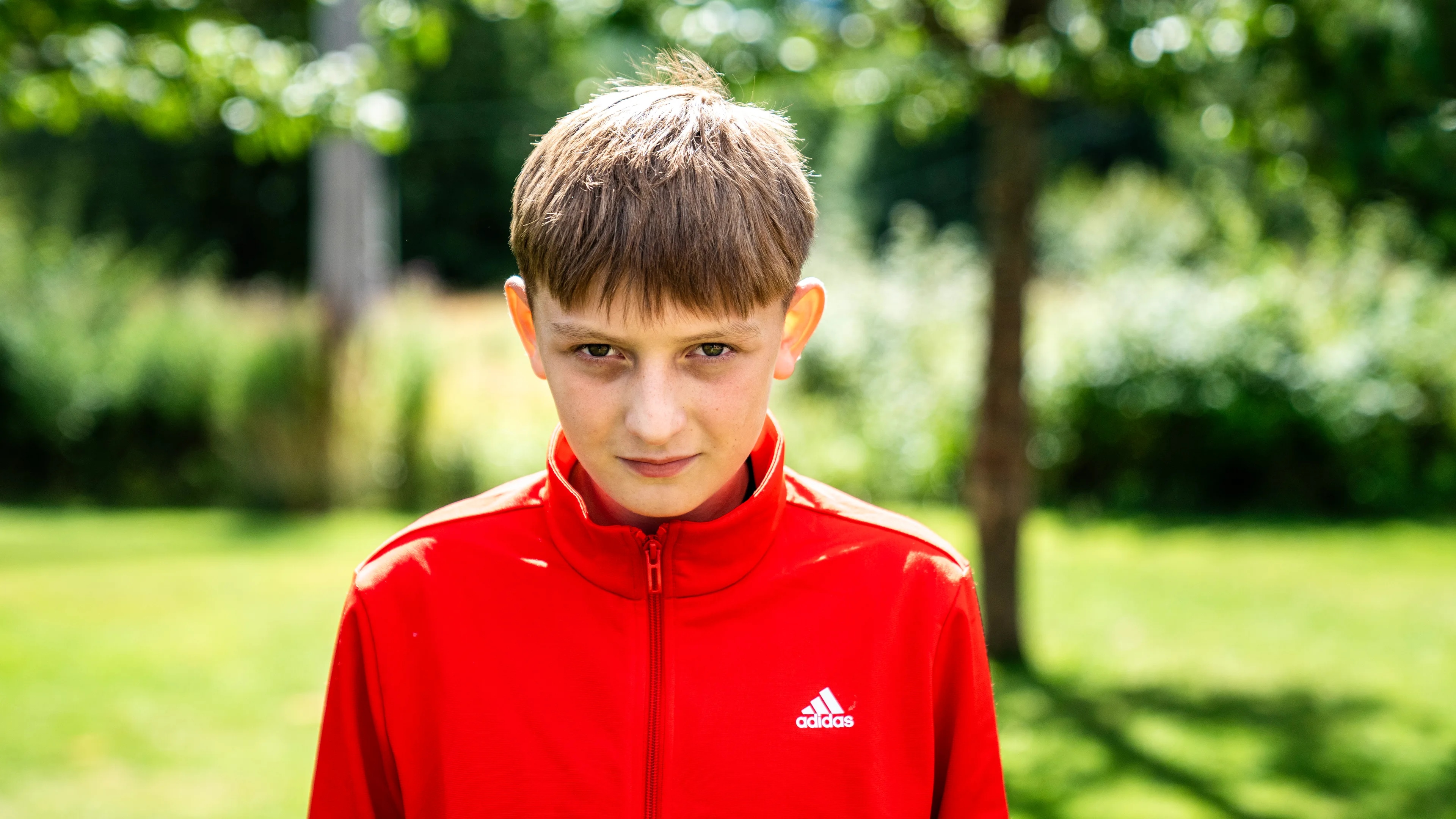Portrett av alvorlig gutt med rød sportsjakke. Skog og grønn plen i bakgrunn