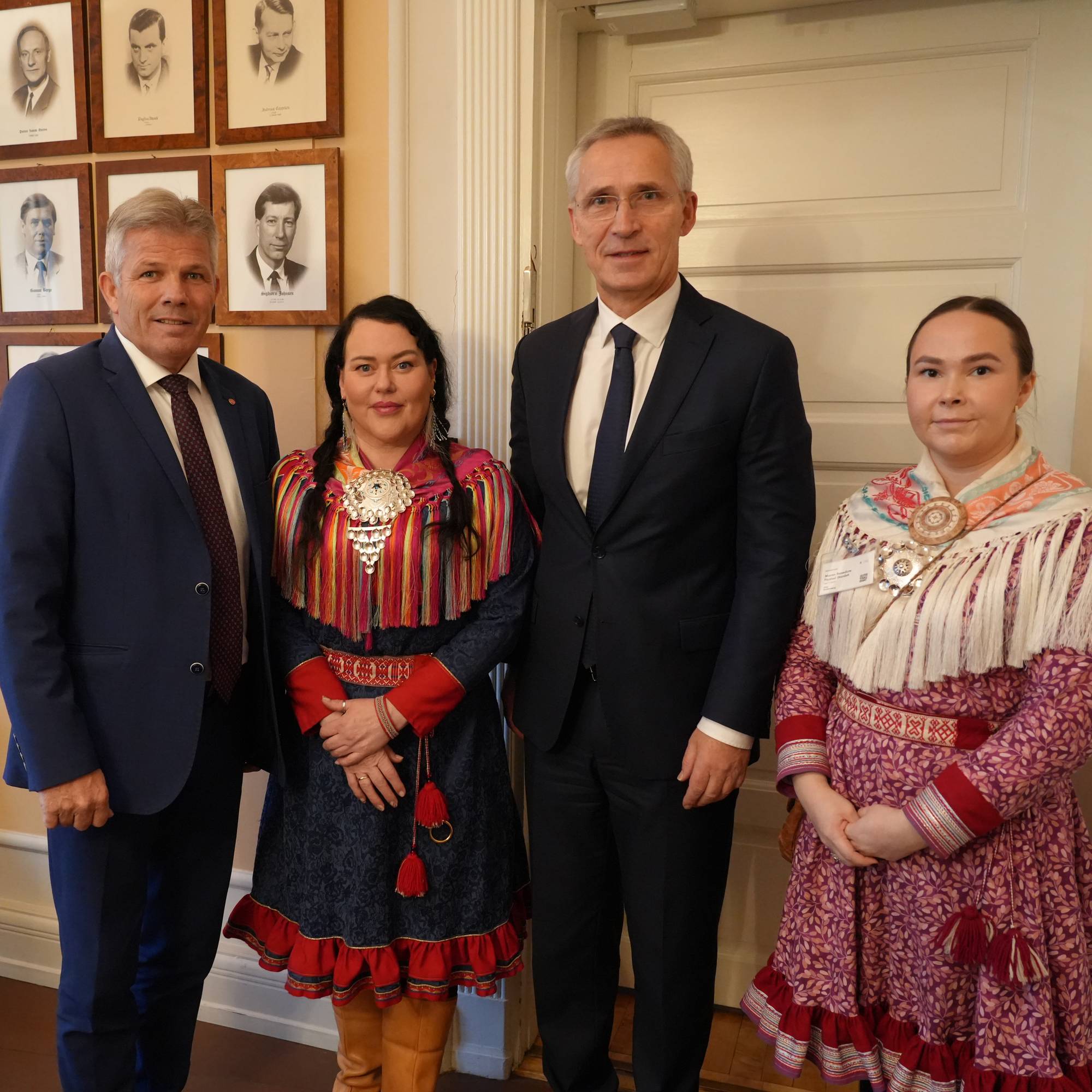 Jens Stoltenberg og tre andre personer står foran en vegg med portrettbilder. (Bildebeskrivelsen er laget av en KI-tjeneste)
