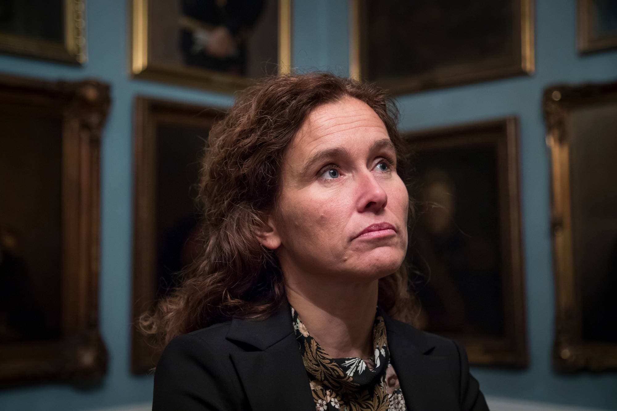 Kvinne med brunt hår i mørk dressjakke står foran bilderammer på en vegg. (Bildebeskrivelsen er laget av en KI-tjeneste)