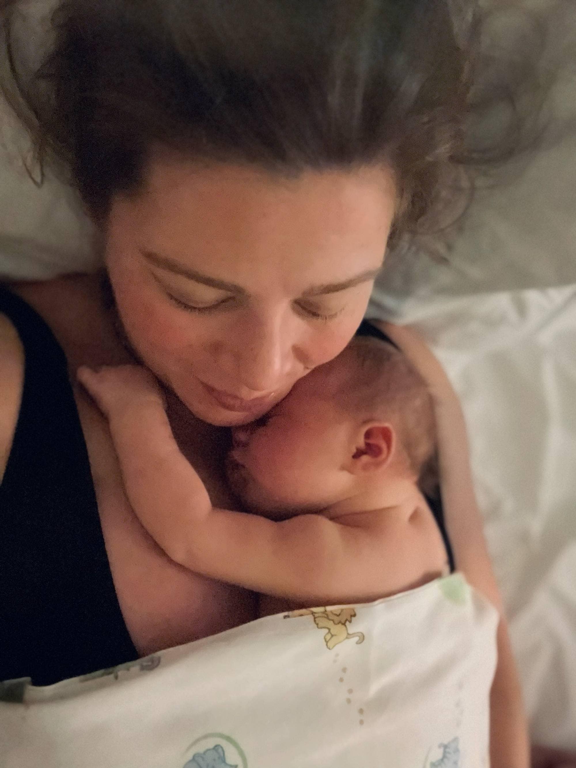 En kvinne ligger i en seng med en nyfødt baby tett inntil brystet. Begge hviler under et teppe. 