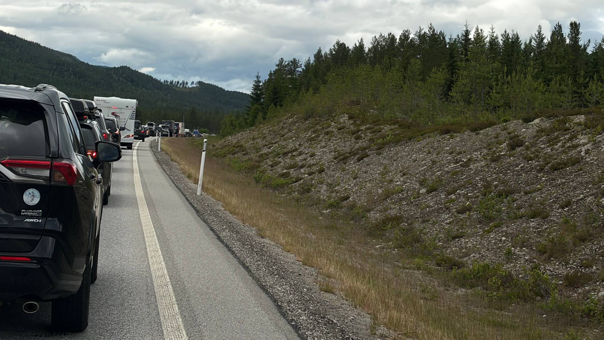 Bilkolonne med biler og campingvogn på en vei med skog og grå himmel i bakgrunnen.