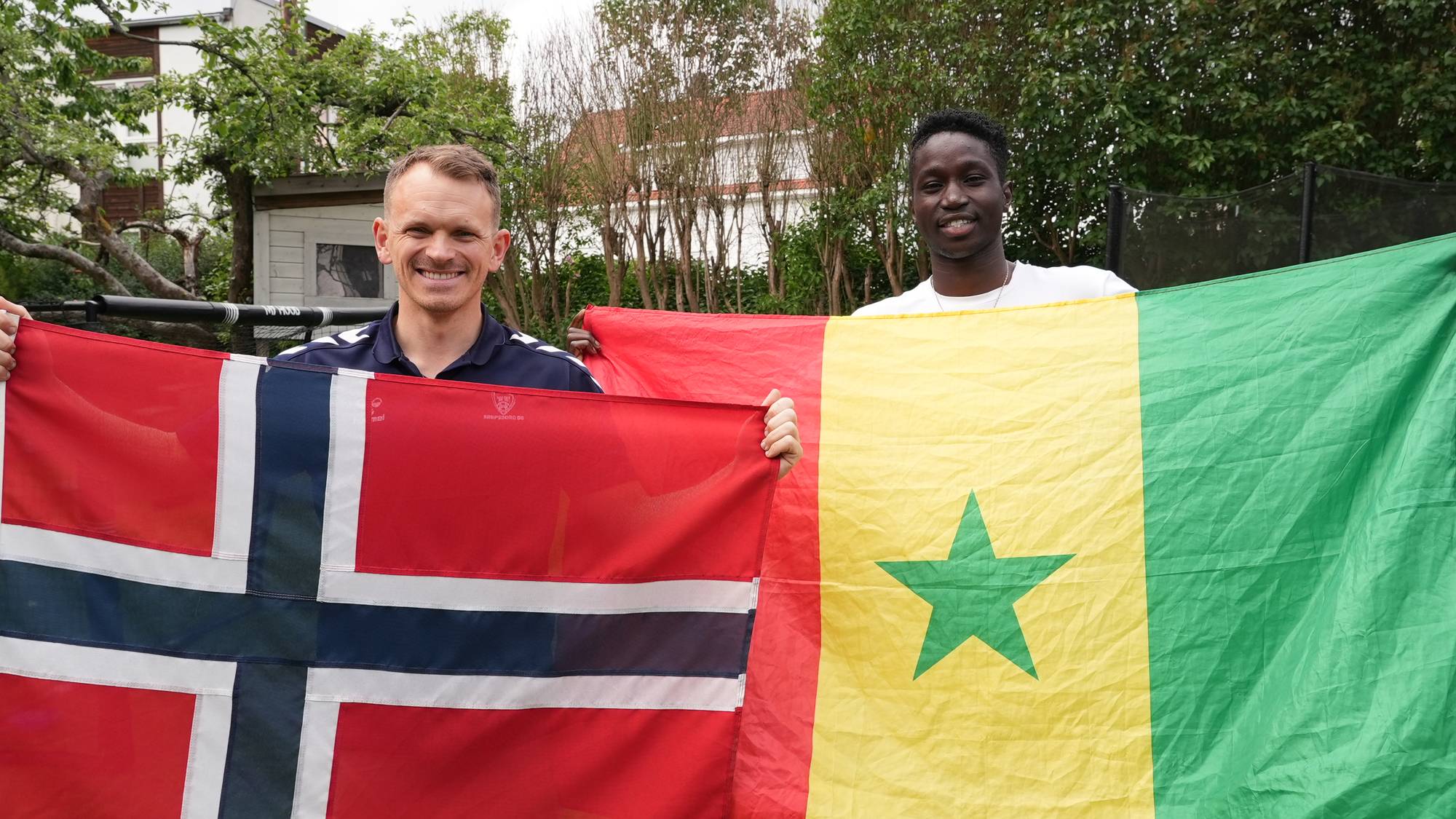 To menn holder hver sin nasjonalflagg. En mann på venstre side holder det norske flagget. Mannen på høyre side holder det senegalesiske flagget. Begge mennene smiler mot kamera. (Bildebeskrivelsen er laget av en KI-tjeneste)