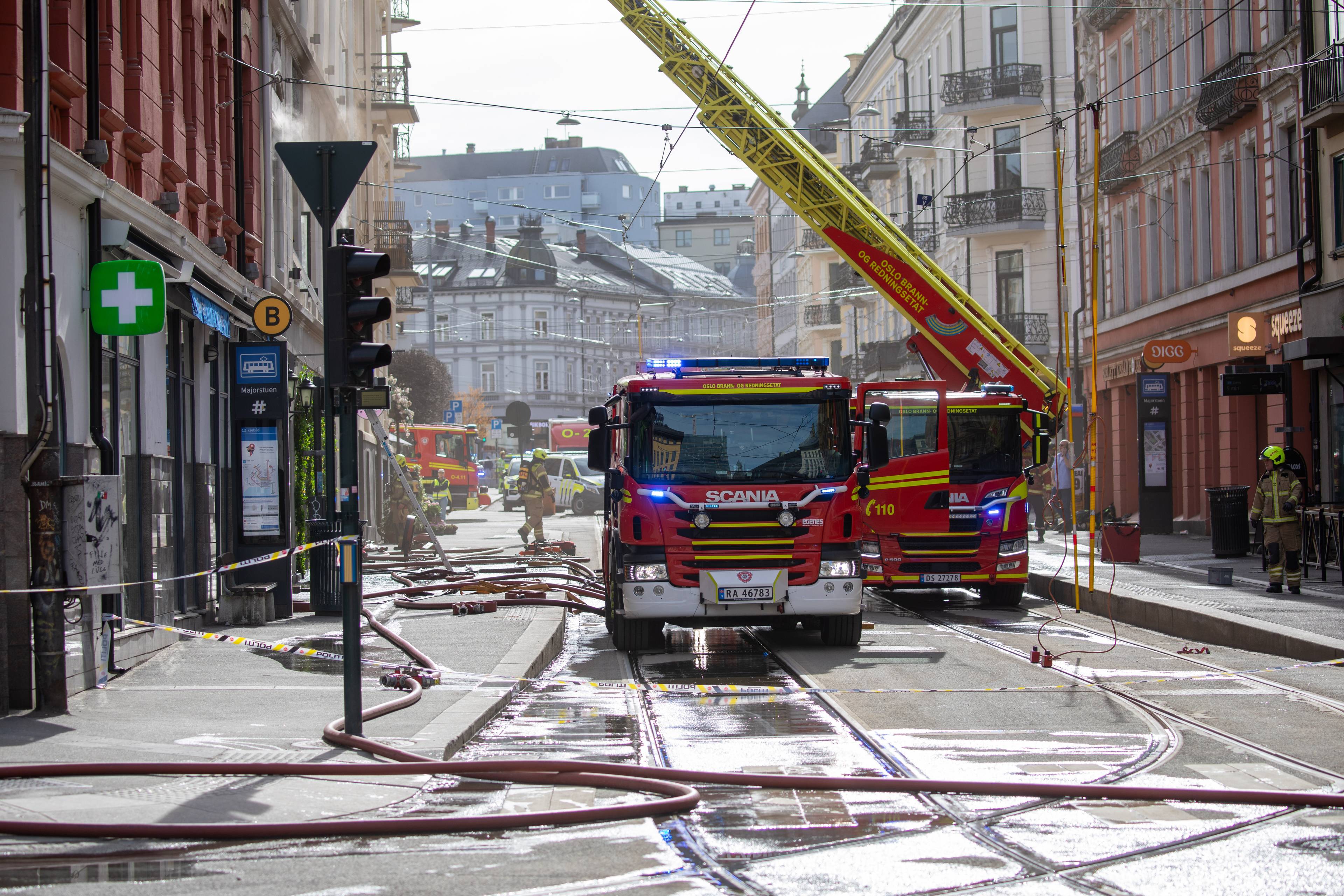 Brannmannskap er på stedet med brannbiler og en lang stige. Det er lagt ut røde slanger på en gate med trikkeskinner. Enkelte bygninger og en trafikklys er synlige i bakgrunnen. (Bildebeskrivelsen er laget av en KI-tjeneste)