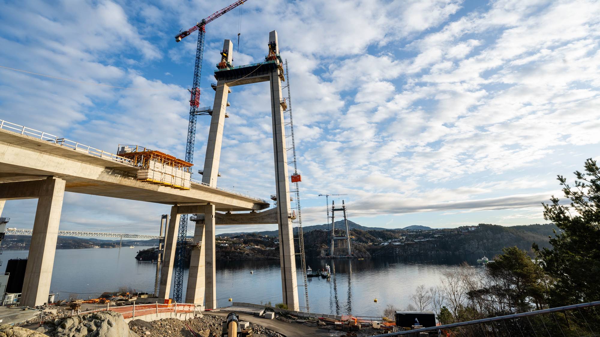 Ene siden av den nye Sotrabruen under konstruksjon. Brotårnene ruver høyt over fjorden. En kran arbeider med å løfte materialer. 