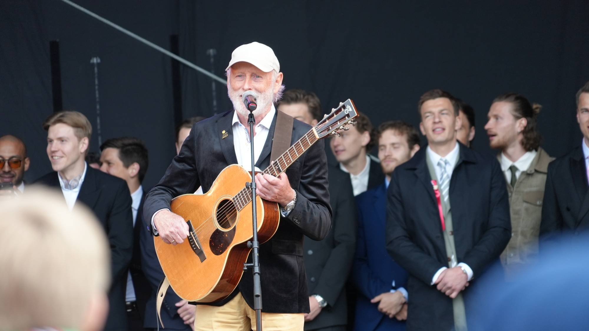 En eldre mann med skjegg spiller gitar og synger på en scene. Han har på seg en hvit caps, en mørk blazer og lyse bukser. Rundt ham står flere menn i dress eller jakke. De ser ut til å være en del av et publikum eller en gruppe. (Bildebeskrivelsen er laget av en KI-tjeneste)