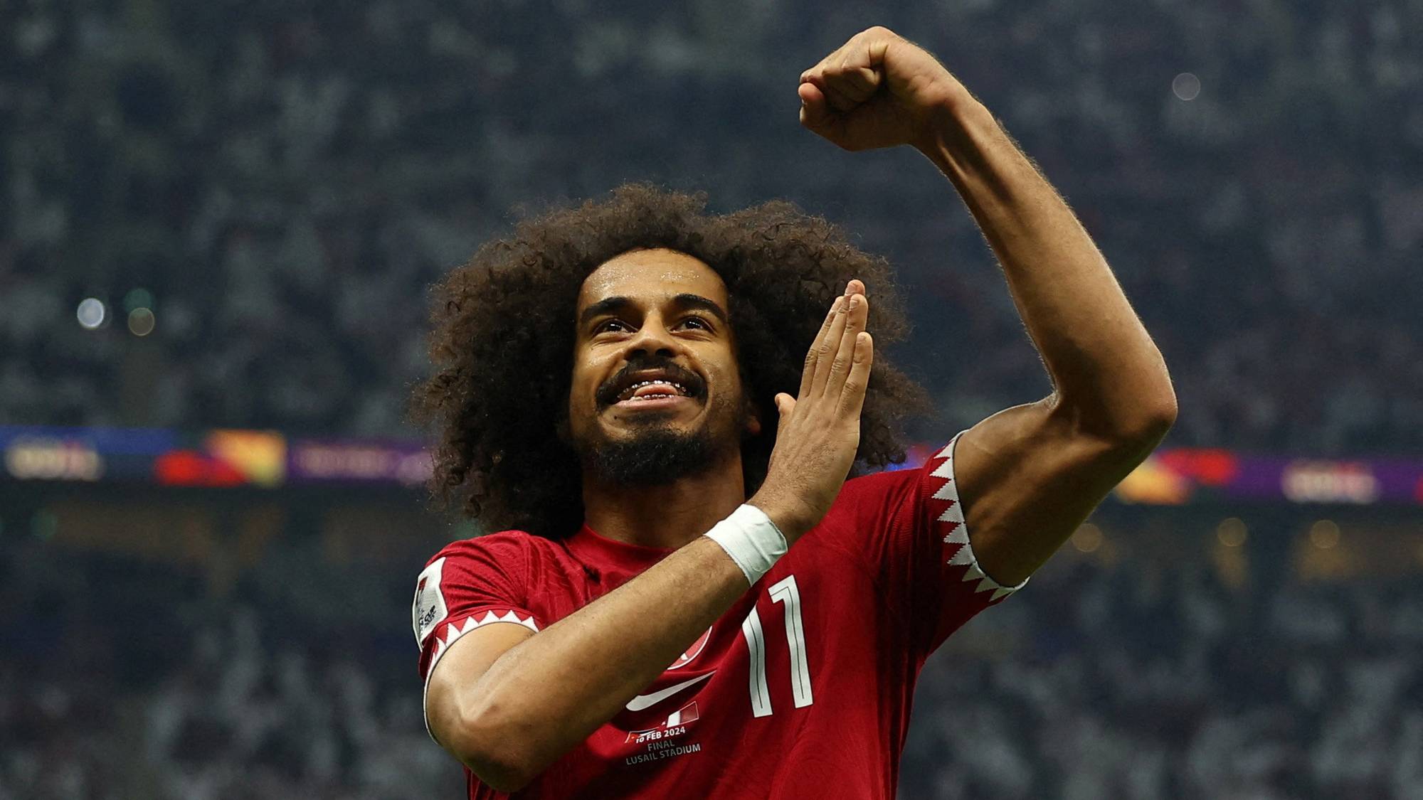 Fotballspiller Akram Afif jubler med armene hevet på Lusail stadion. Han har på seg en rød drakt med nummer 11. I bakgrunnen er det tilskuere. (Bildebeskrivelsen er laget av en KI-tjeneste)