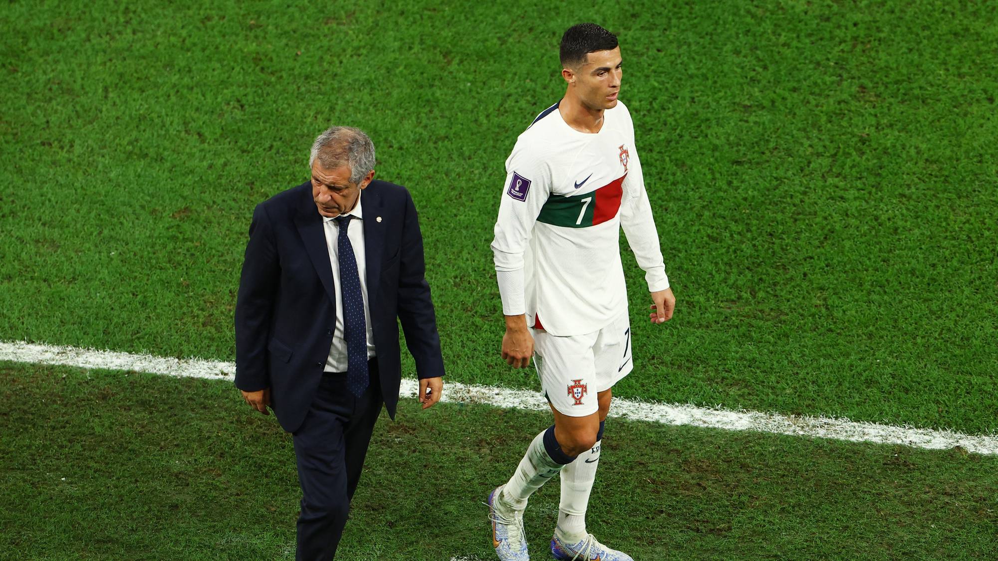 Fotballspiller Cristiano Ronaldo og landslagstrener Fernando Santos går på en fotballbane. Ronaldo er iført en hvit drakt med nummer 7. Santos er iført mørk dress. (Bildebeskrivelsen er laget av en KI-tjeneste)