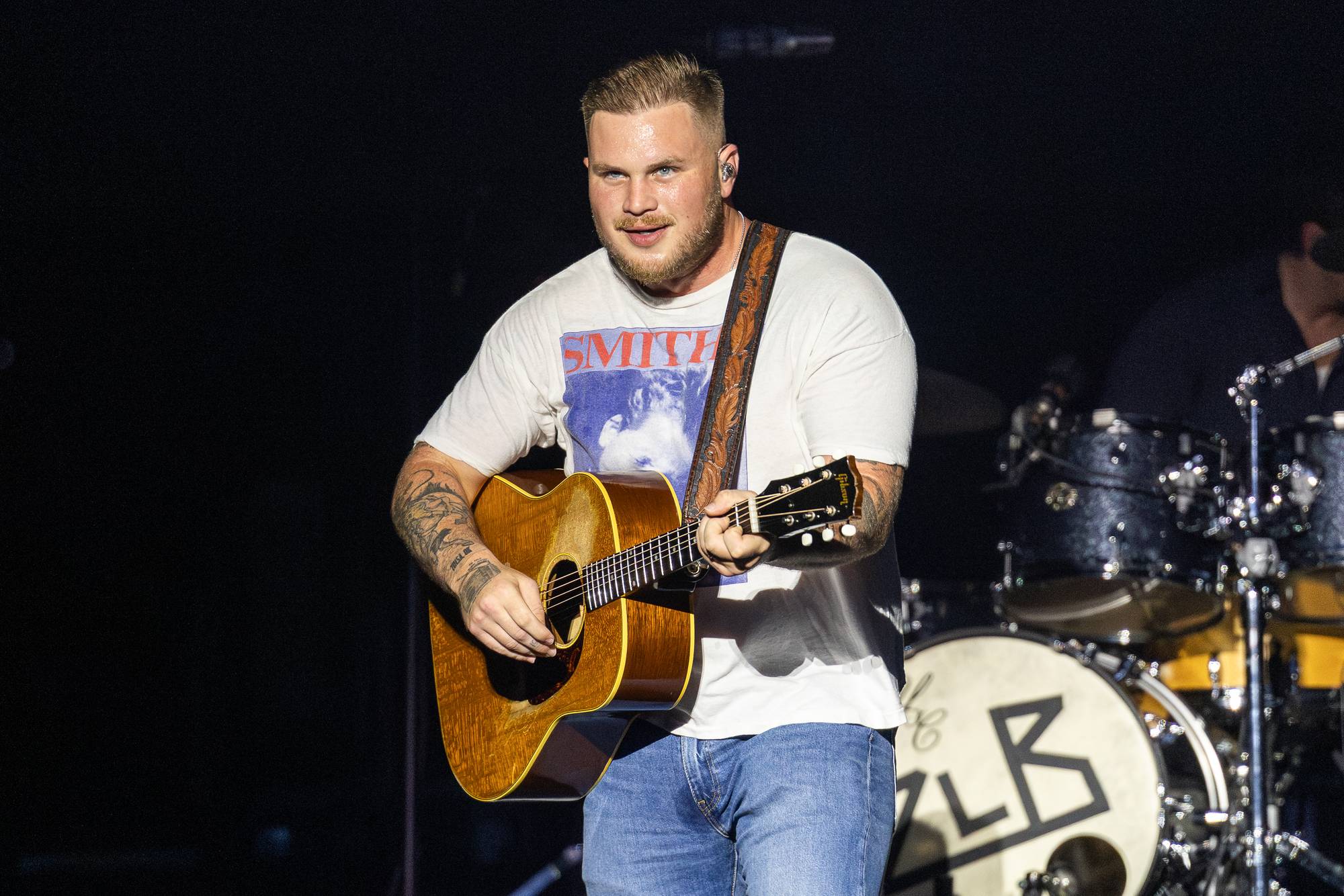 En mann med gitar spiller på en scene. Han har på seg en t-skjorte med trykk og blå jeans. Bak ham er et trommesett. (Bildebeskrivelsen er laget av en KI-tjeneste)