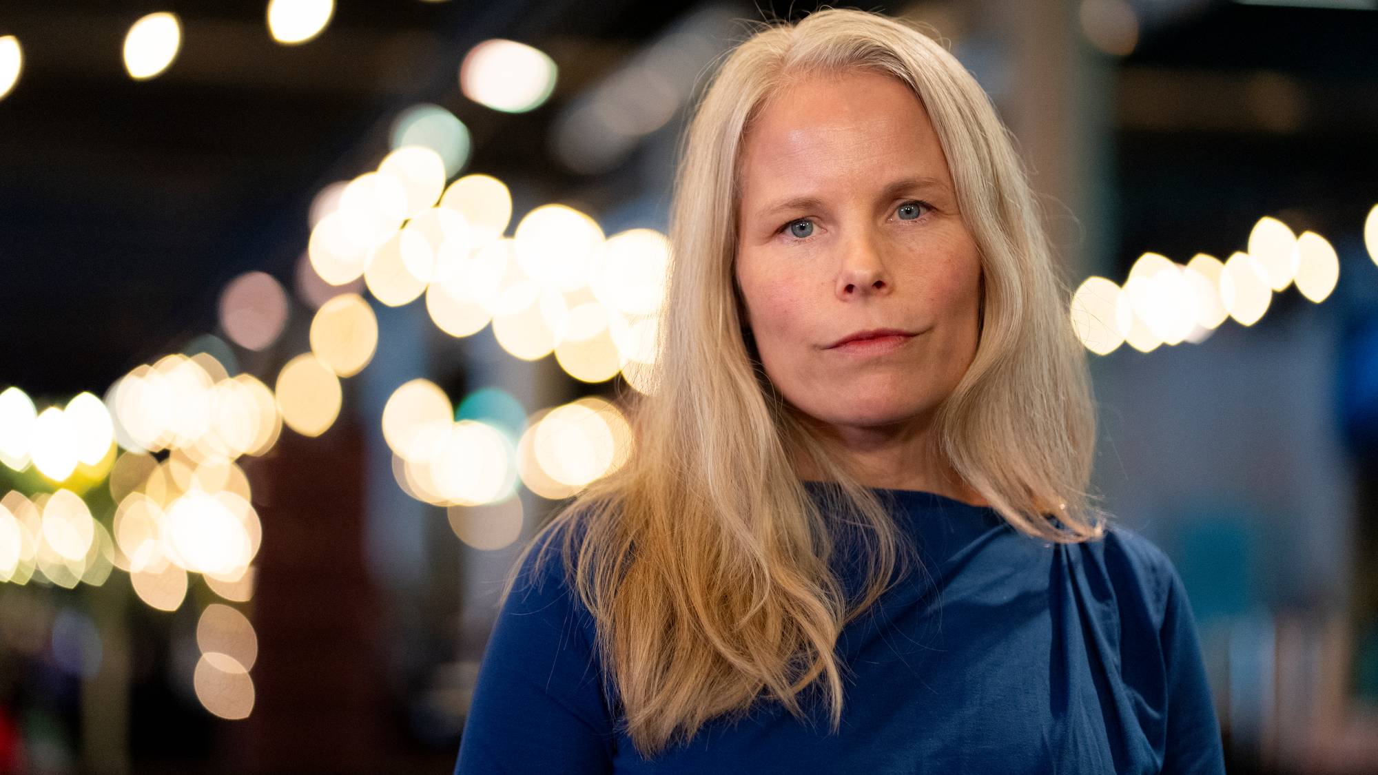 En kvinne med langt, lyst hår står foran et bakgrunnsbelyst område med uklare lys. Hun har på seg en blå genser og ser mot kameraet. Omgivelsene gir en uformell atmosfære, delvis med flere lysørd som skaper en hjemmekoselig stemning. Bildet er tatt innendørs, muligens i en restaurant eller et arrangementslokale. (Bildebeskrivelsen er laget av en KI-tjeneste)