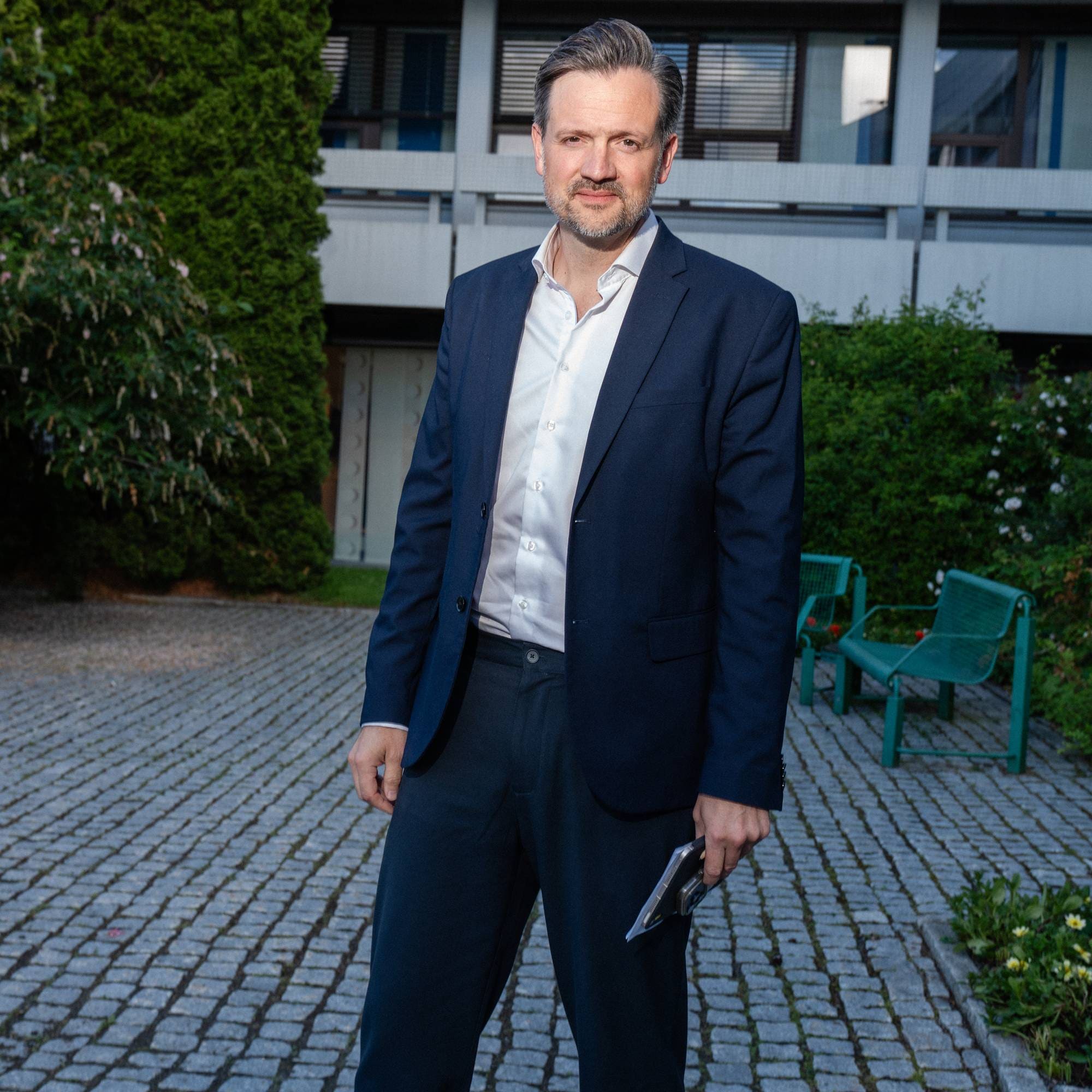 Mann i mørk dress og hvit skjorte står ute. Han holder en telefon i høyre hånd. Bak ham er det en grønn busk og en bygning. Han står på et brosteinsbelagt underlag. (Bildebeskrivelsen er laget av en KI-tjeneste)