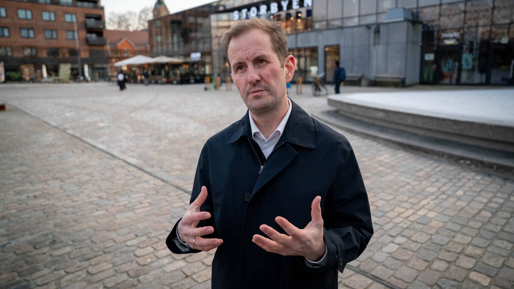 En mann snakker med utstrakte hender på en brosteinsbelagt plass. Bakgrunnen viser bygninger og kafeer. Det er få mennesker til stede i området. En kirketårn er synlig i det fjerne.