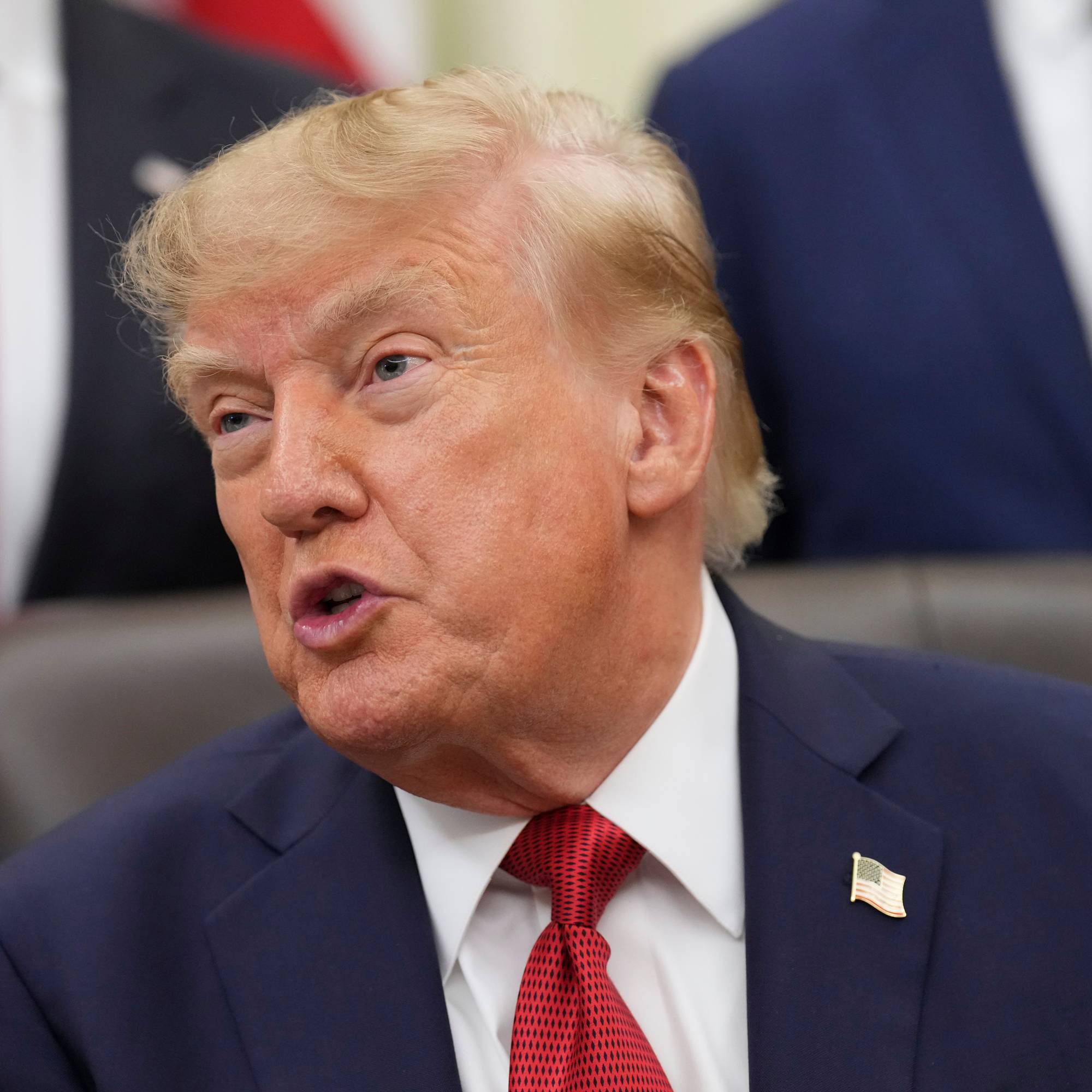 Donald Trump i dress og slips snakker under et møte. Personer i bakgrunnen er delvis synlige. (Bildebeskrivelsen er laget av en KI-tjeneste)