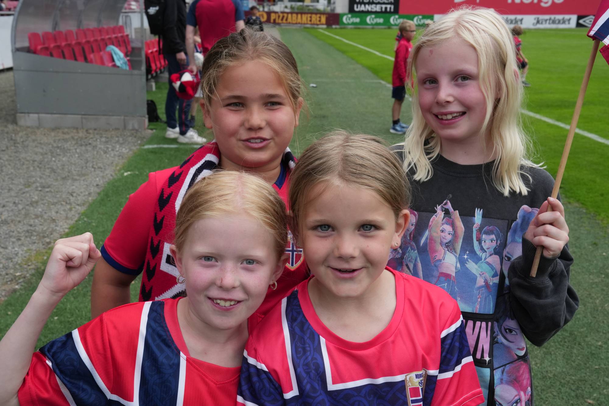 Fire jenter står i fotballdrakter på en fotballbane og smiler. 