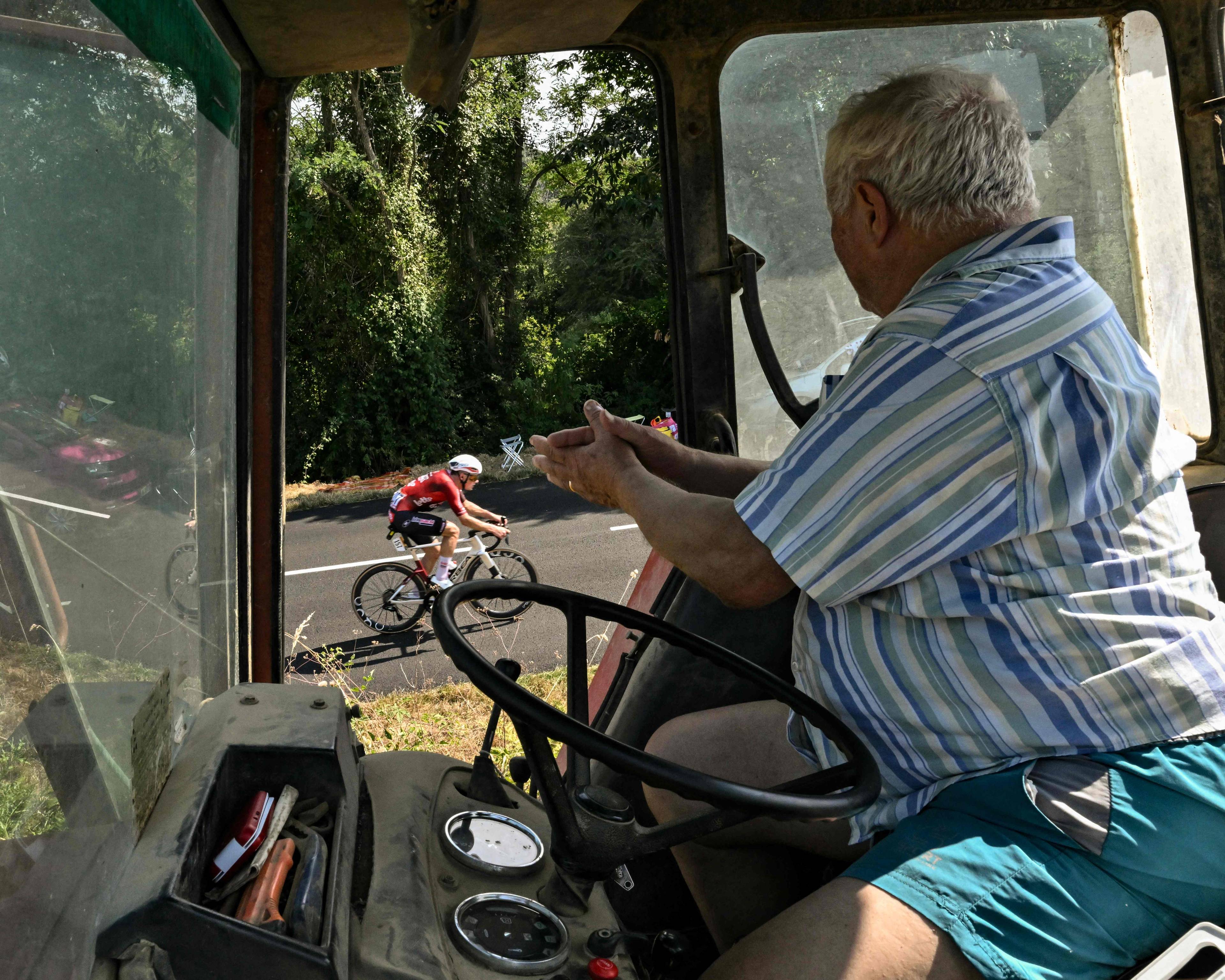 En eldre mann sitter i førerhuset på en traktor og ser på en syklist som passerer på veien. (Bildebeskrivelsen er laget av en KI-tjeneste)