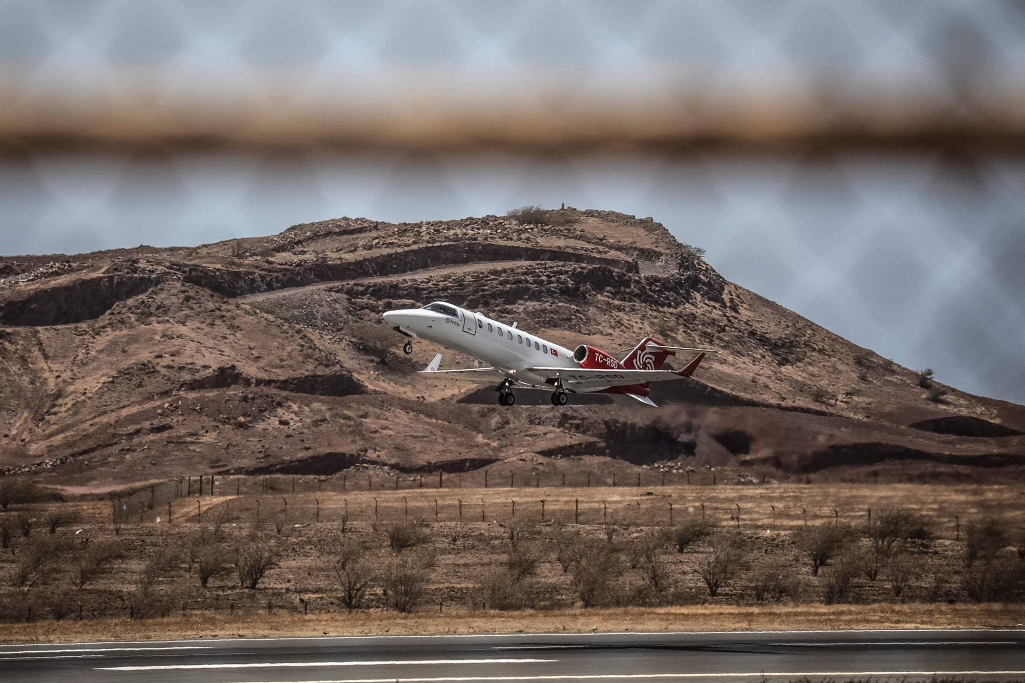 Et privatfly letter fra en rullebane. Flyet er hvitt med røde detaljer og har registreringen TC-RSD. I bakgrunnen er et goldt fjellandskap. Bildet er tatt gjennom et gjerde. (Bildebeskrivelsen er laget av en KI-tjeneste)