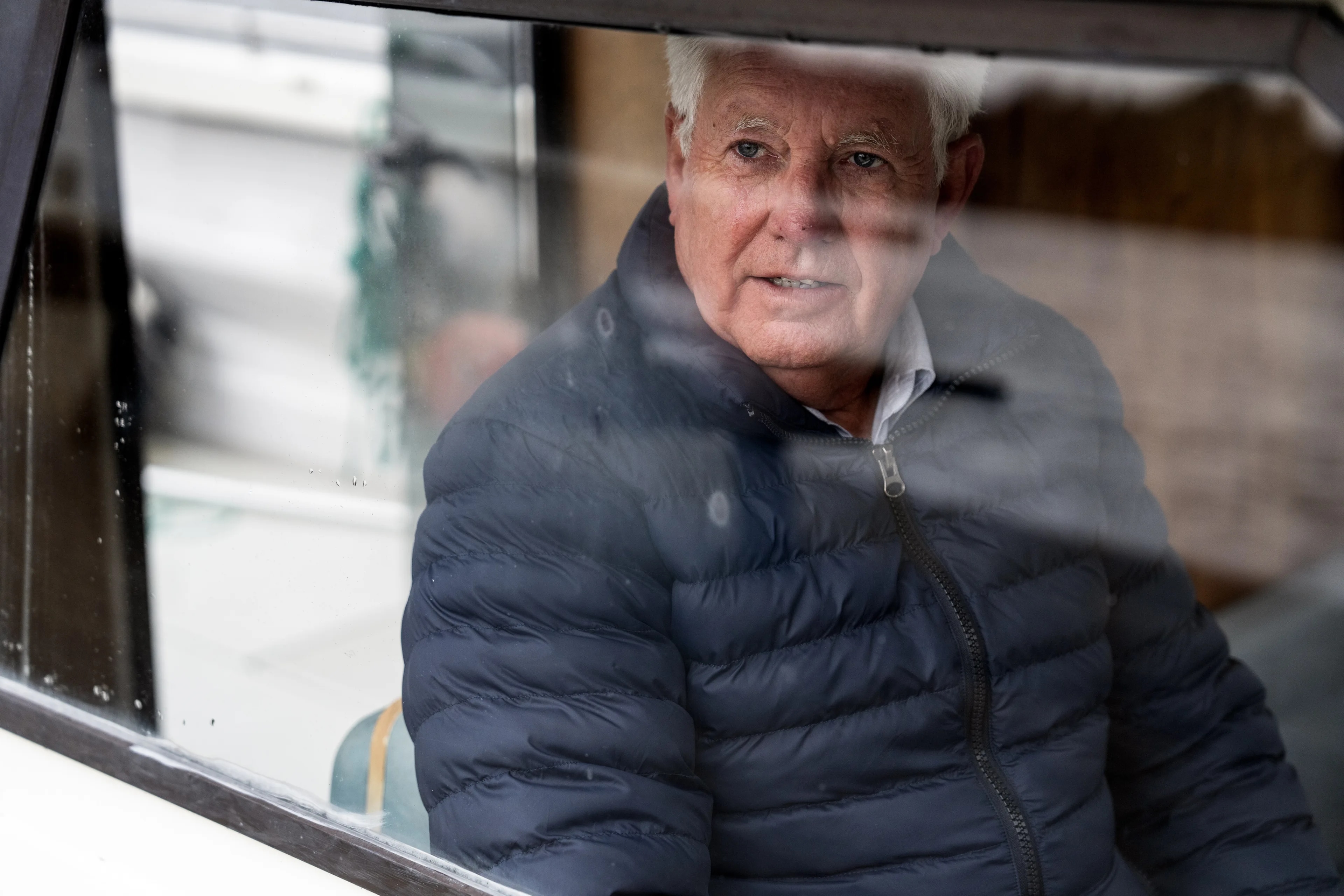 Erik Arntzen sitter med blå boblejakke bak et vindu, og ser ut til siden. Han har hvitt hår og et alvorlig ansiktsutrykk.