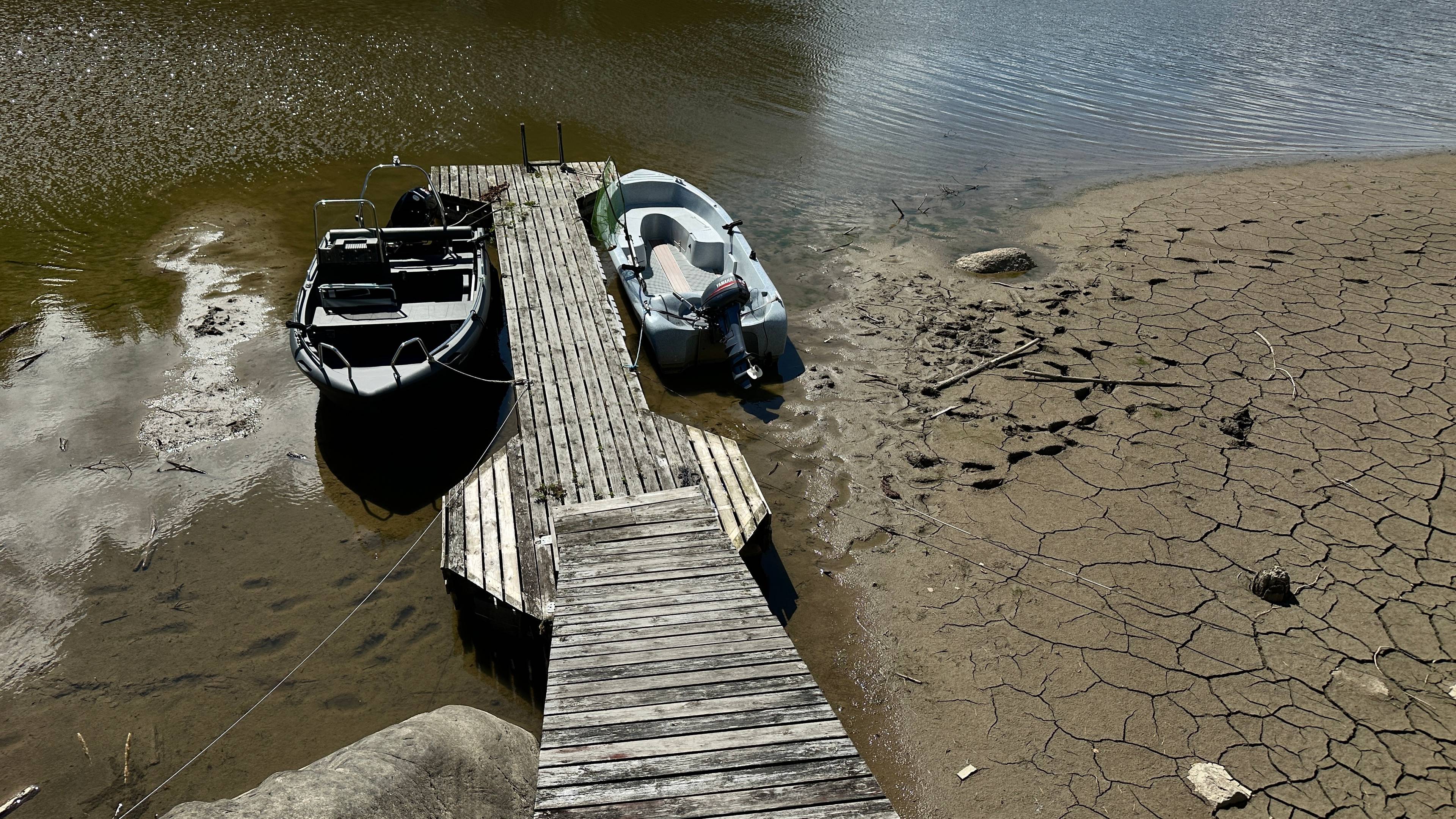 To småbåter ligger fortøyd ved en trebrygge omgitt av uttørket og sprukket jordbunn. (Bildebeskrivelsen er laget av en KI-tjeneste)