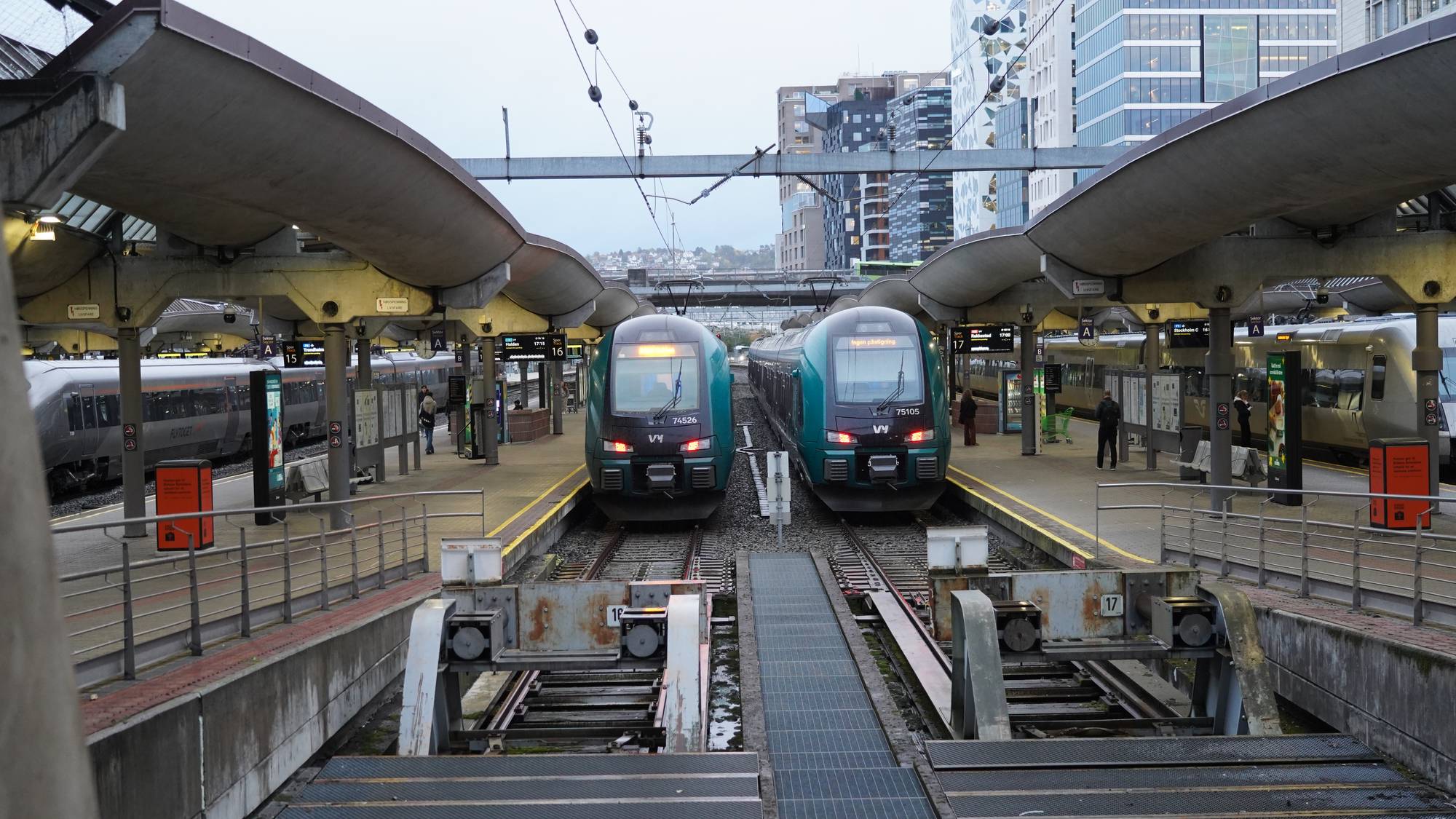 To Vy-tog står ved plattformene på Oslo S. Moderne kontorbygg i bakgrunnen. (Bildebeskrivelsen er laget av en KI-tjeneste)