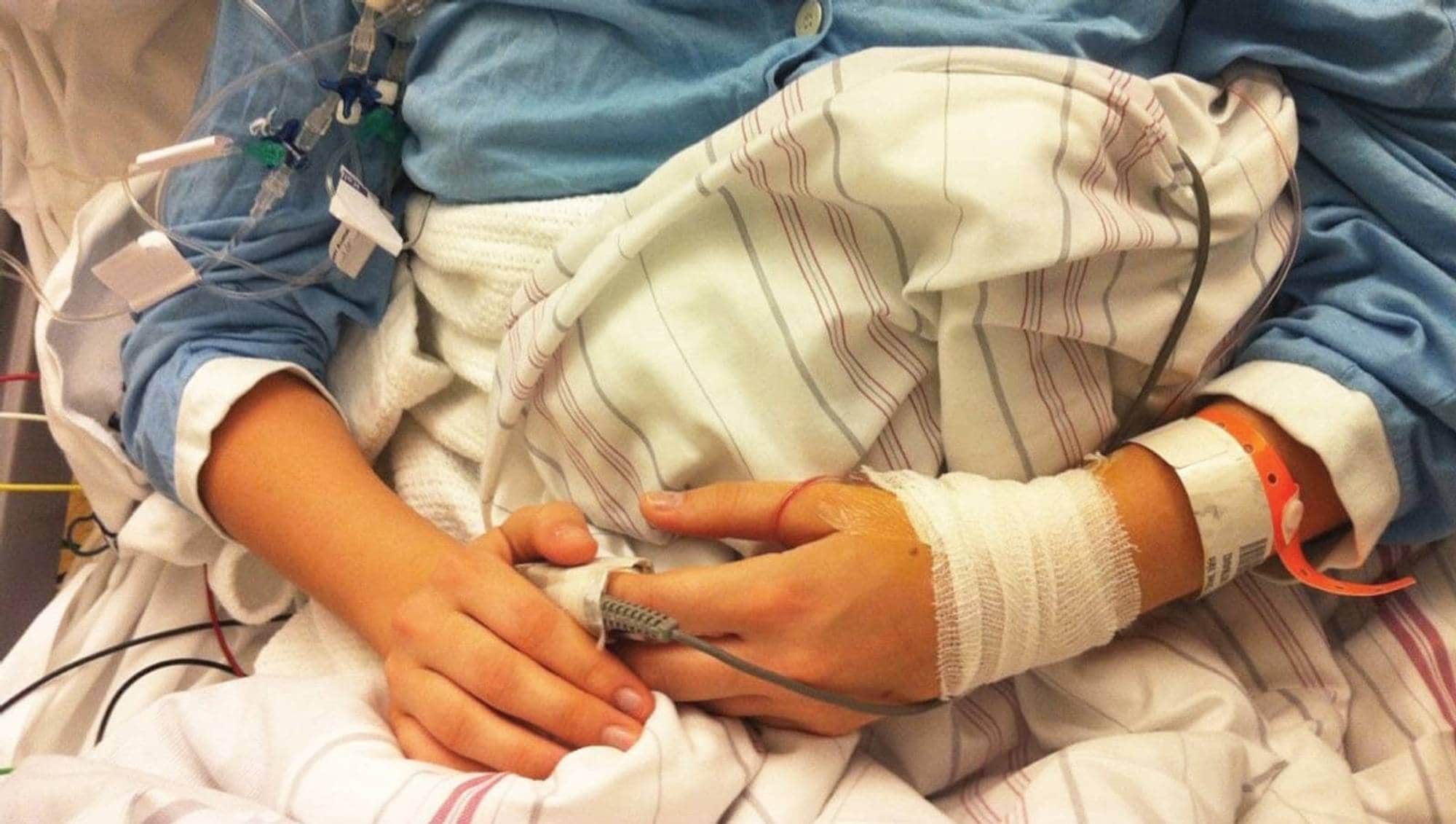 En person ligger i en sykehusseng med medisinsk utstyr. En pulsoksymeter er festet til en finger. Personen har intravenøs tilgang og bandasje på armen. Et pasientarmbånd er synlig. (Bildebeskrivelsen er laget av en KI-tjeneste)