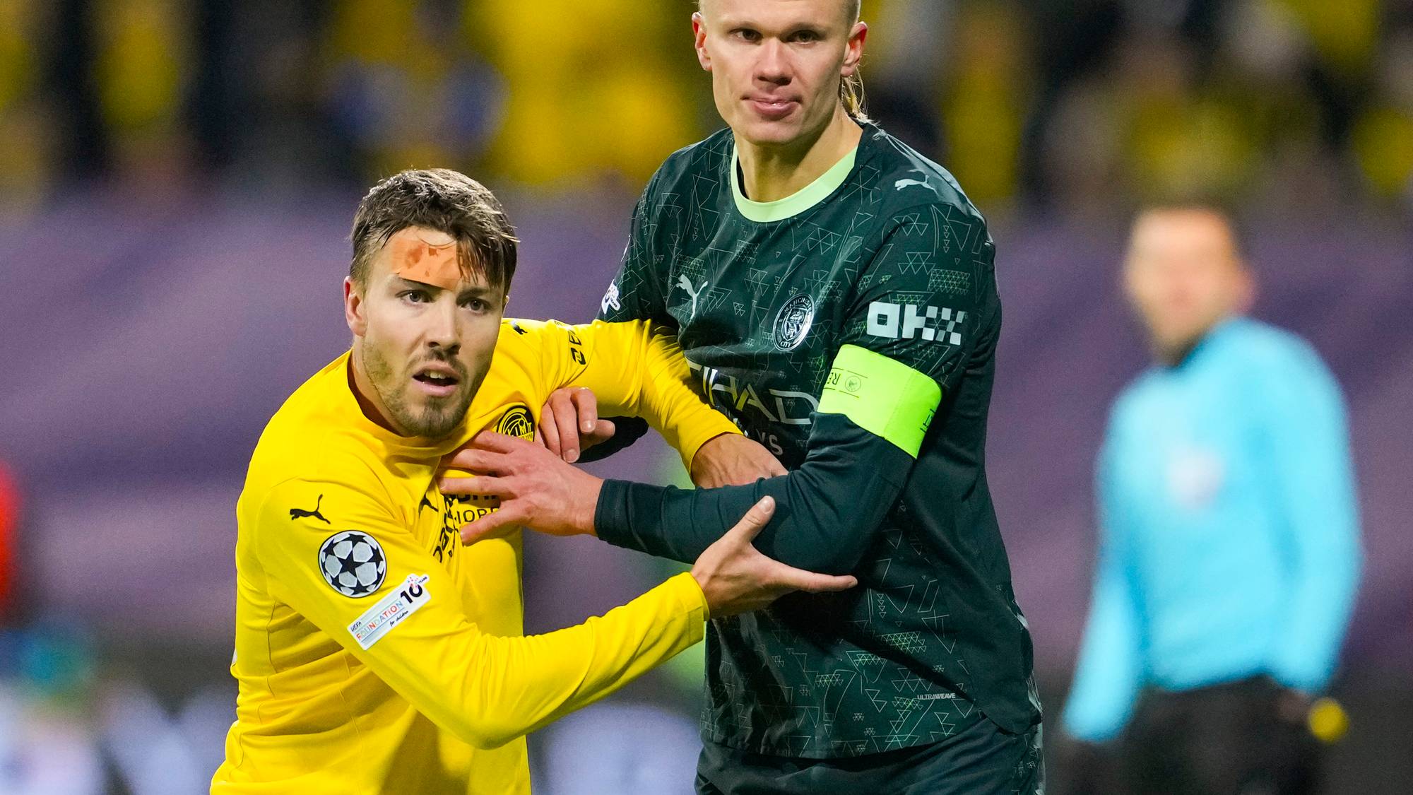 To personer i fotballuniformer kjemper om ballen på en fotballbane. Den ene spilleren har på seg en gul drakt, mens den andre har på seg en grønn og svart drakt med kapteinsbånd. Publikum i bakgrunnen vises med gule klær, noe som tyder på en hjemmekamp. Dommeren står i bakgrunnen og observerer situasjonen.