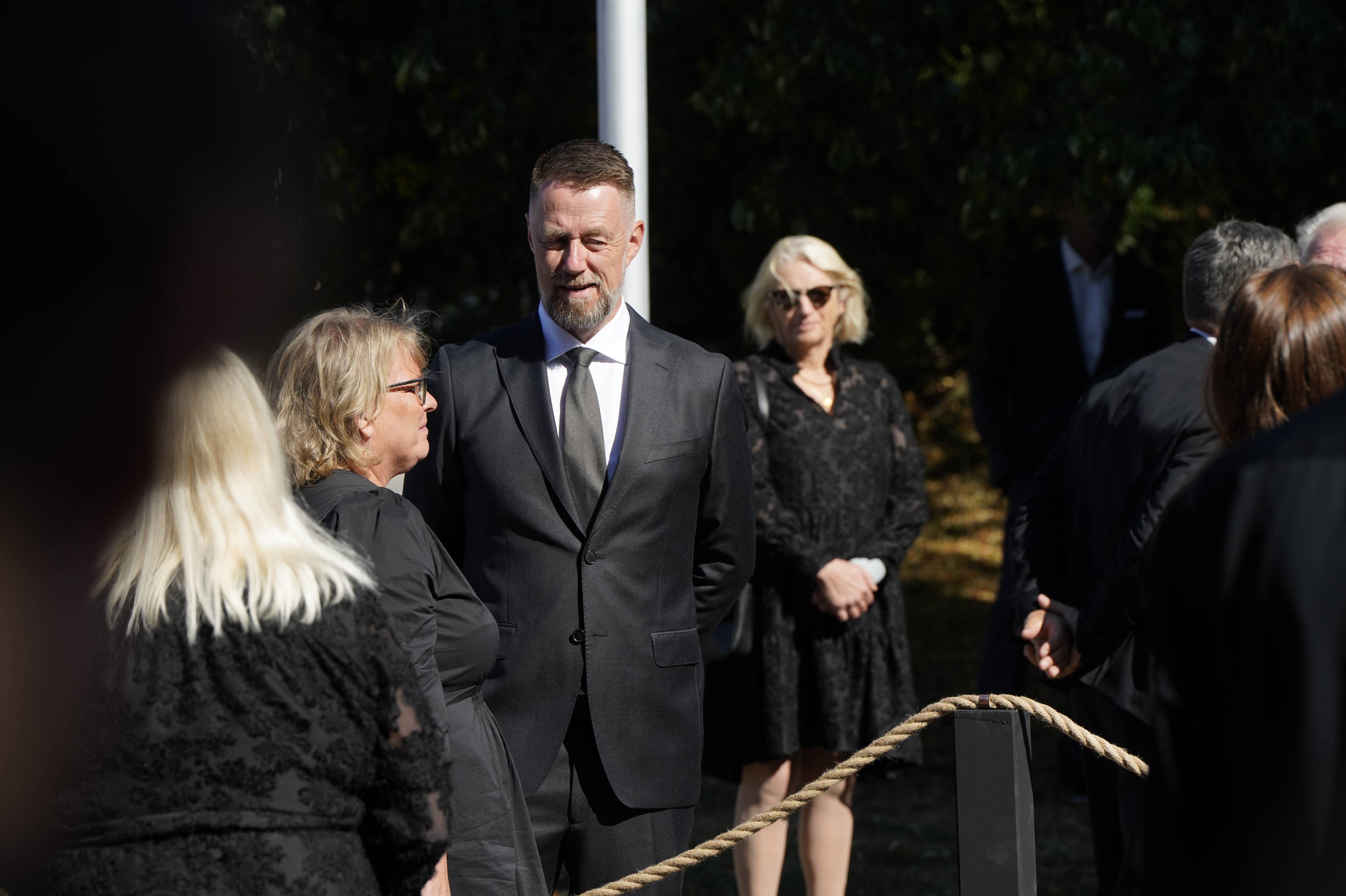 En mann i mørk dress står sammen med flere personer kledd i svart utendørs.