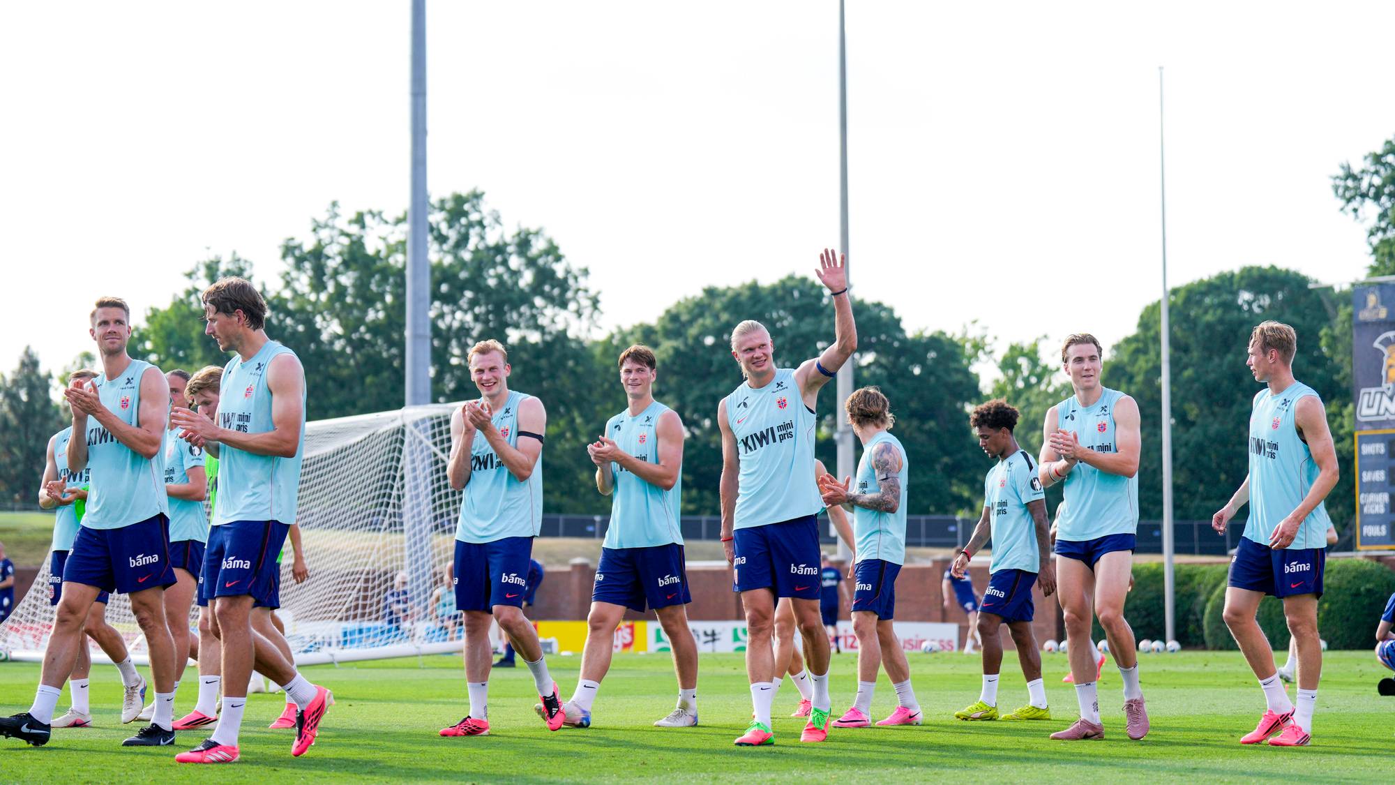 En gruppe menn i lyseblå fotballtrøyer og mørkeblå shorts trener på en gressbane. Én av mennene vinker til kameraet. Mennene har på seg fotballsko og flere av dem klapper. Bak mennene er det et fotballmål og trær. (Bildebeskrivelsen er laget av en KI-tjeneste)