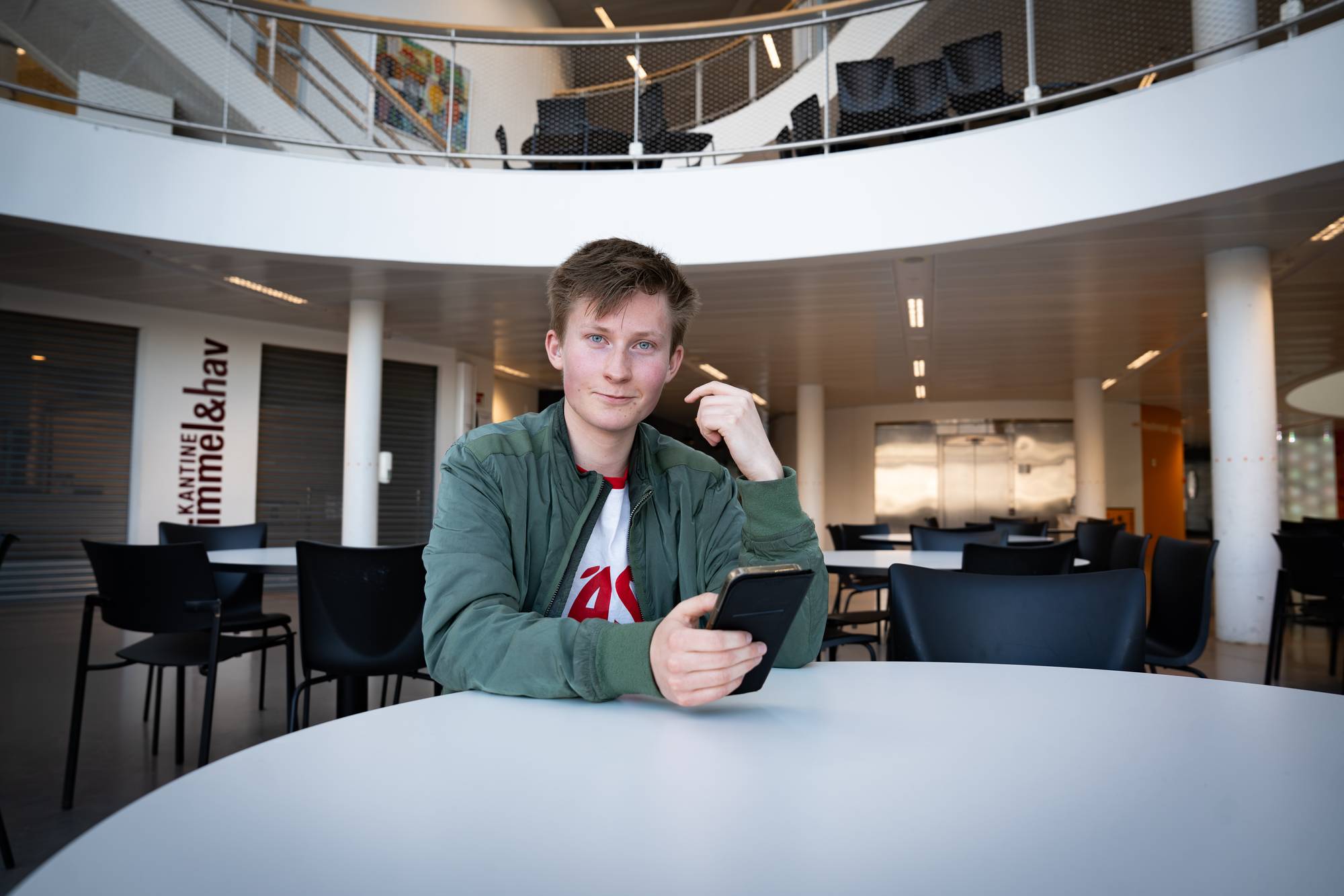 Christian sitter ved et rundt bord og ser på mobilen. Han har på seg en grønn jakke over en hvit t-skjorte. Bak ham er det et stort, åpent rom med bord og stoler. En tekst på veggen viser "KANTINE Himmel&hav". 