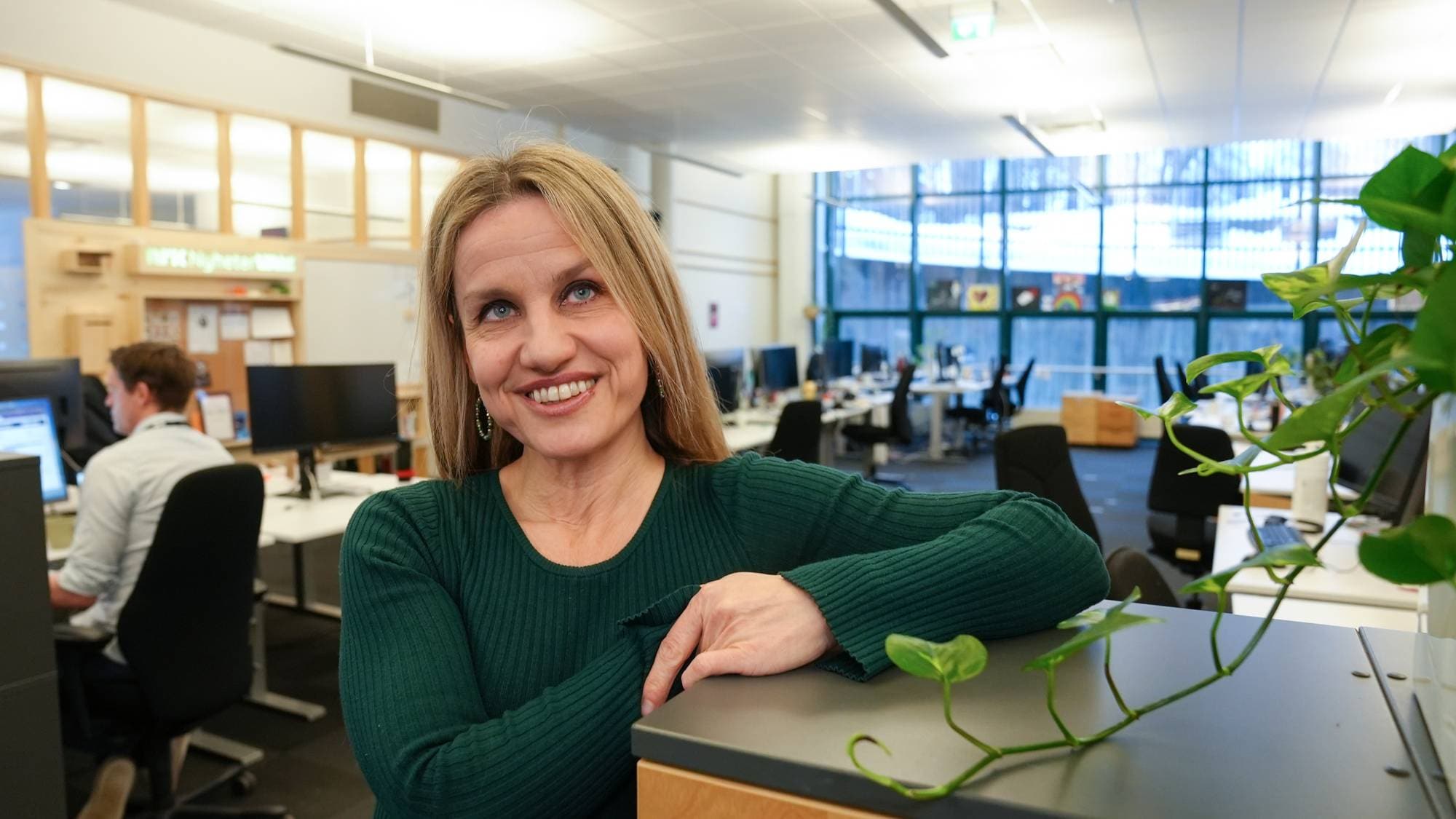 En kvinne i grønt står i et redaksjonslokale og smiler mens hun ser på noen bak kamera.