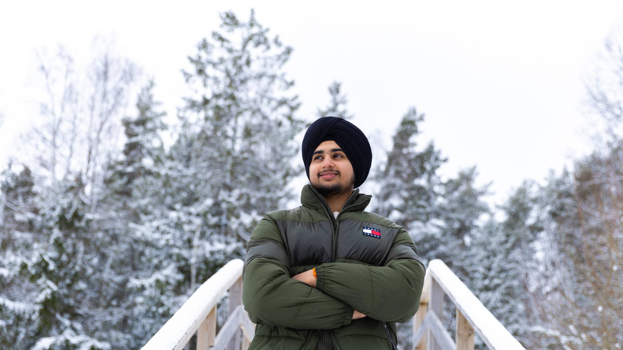 Gurveer står ute i en trapp med armene i kors. Han ser til siden og smiler stolt. Han har på seg grønn jakke og sort turban. Det er snø og vinter ute.