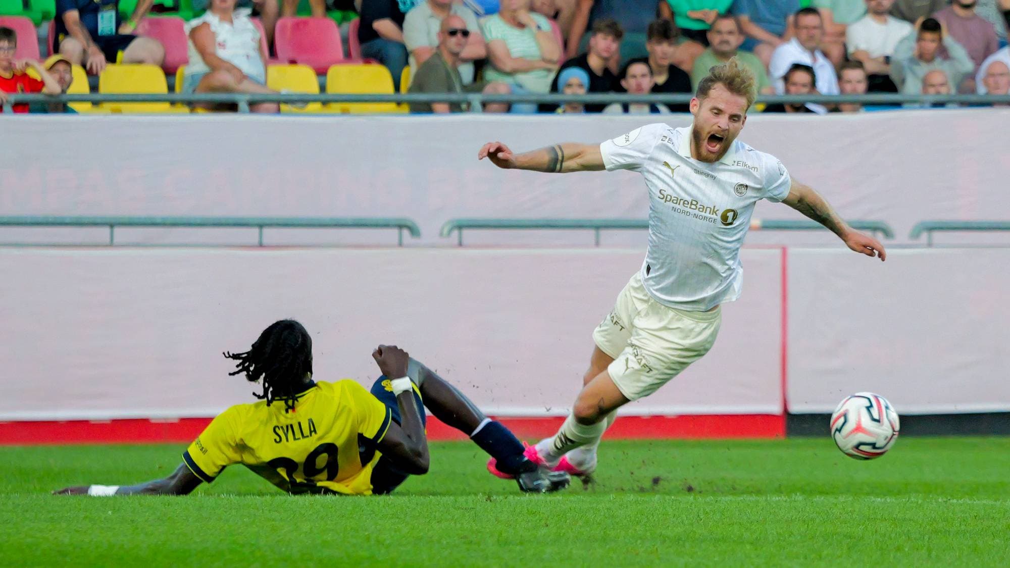En fotballspiller i hvitt går i bakken etter en takling fra en spiller i gult og svart. (Bildebeskrivelsen er laget av en KI-tjeneste)