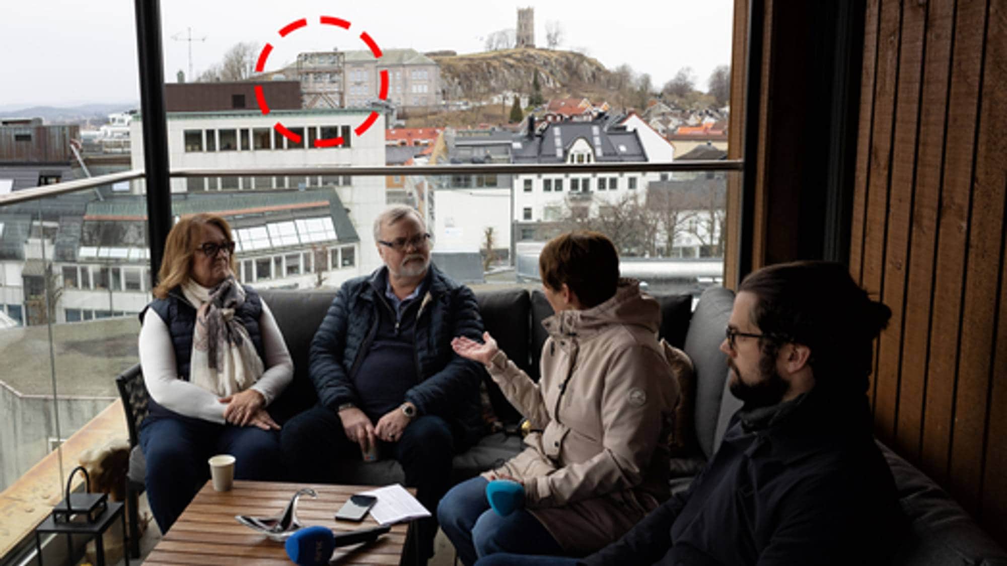 Flere sitter i en sofa rundt et bord. De sitter på en balkong med utsikt over byen. Klokketårnet er markert med en rød ring. 