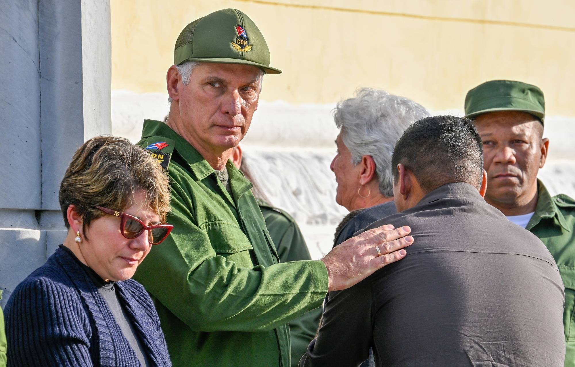 En mann i grønn uniform og med caps med et cubansk emblem legger en hånd på skulderen til en annen mann. En kvinne med solbriller står ved siden av dem. En annen mann i grønn uniform står lengre bak. (Bildebeskrivelsen er laget av en KI-tjeneste)