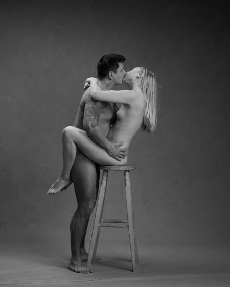En naken dame sitter på en krakk mens hun kysser en naken mann med tatoveringer som står mellom bena hennes