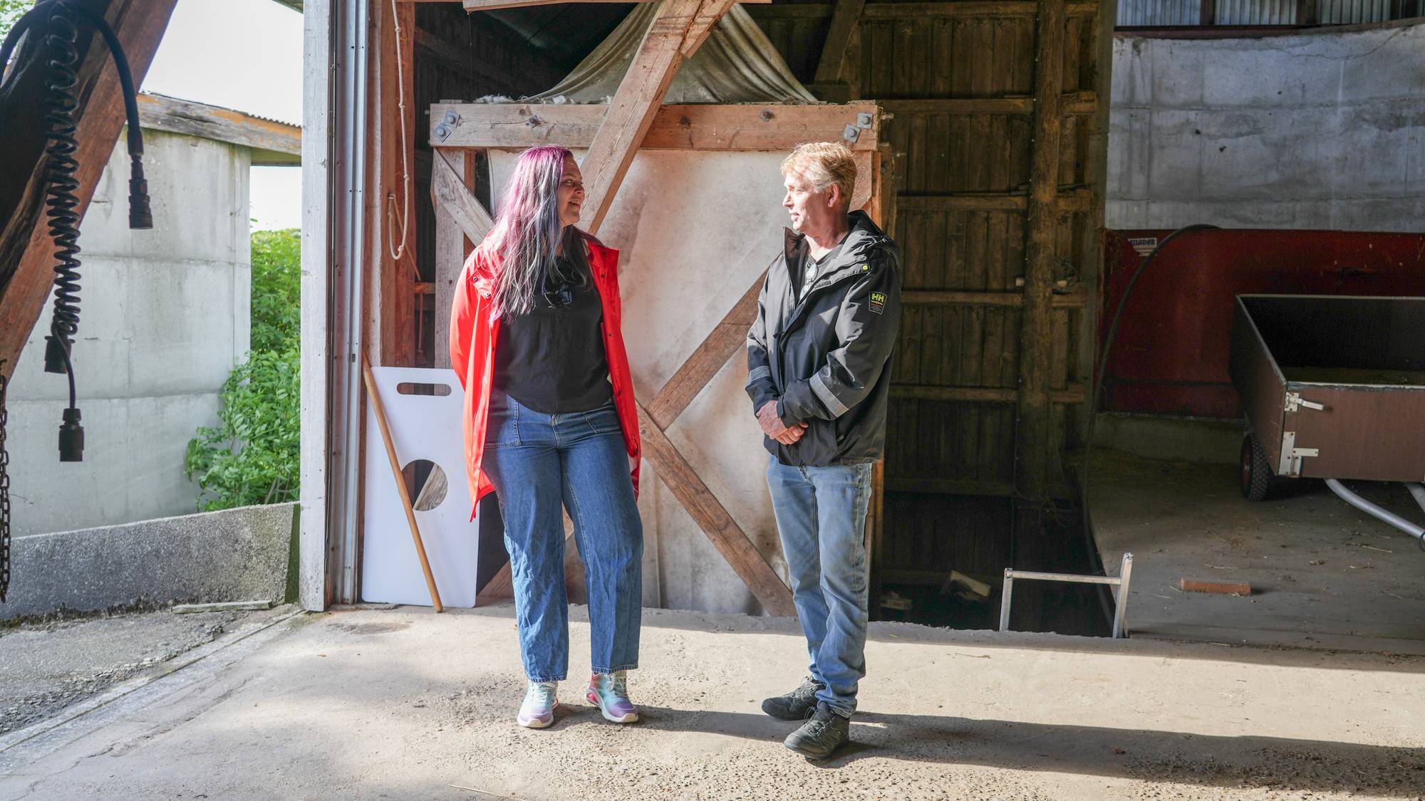 To personer står og snakker sammen inne i en fjøsbygning. Kvinnen til venstre har lilla hår og en rød jakke. Mannen til høyre har på seg en mørk jakke. I bakgrunnen sees en vogn og deler av bygningens konstruksjon. (Bildebeskrivelsen er laget av en KI-tjeneste)