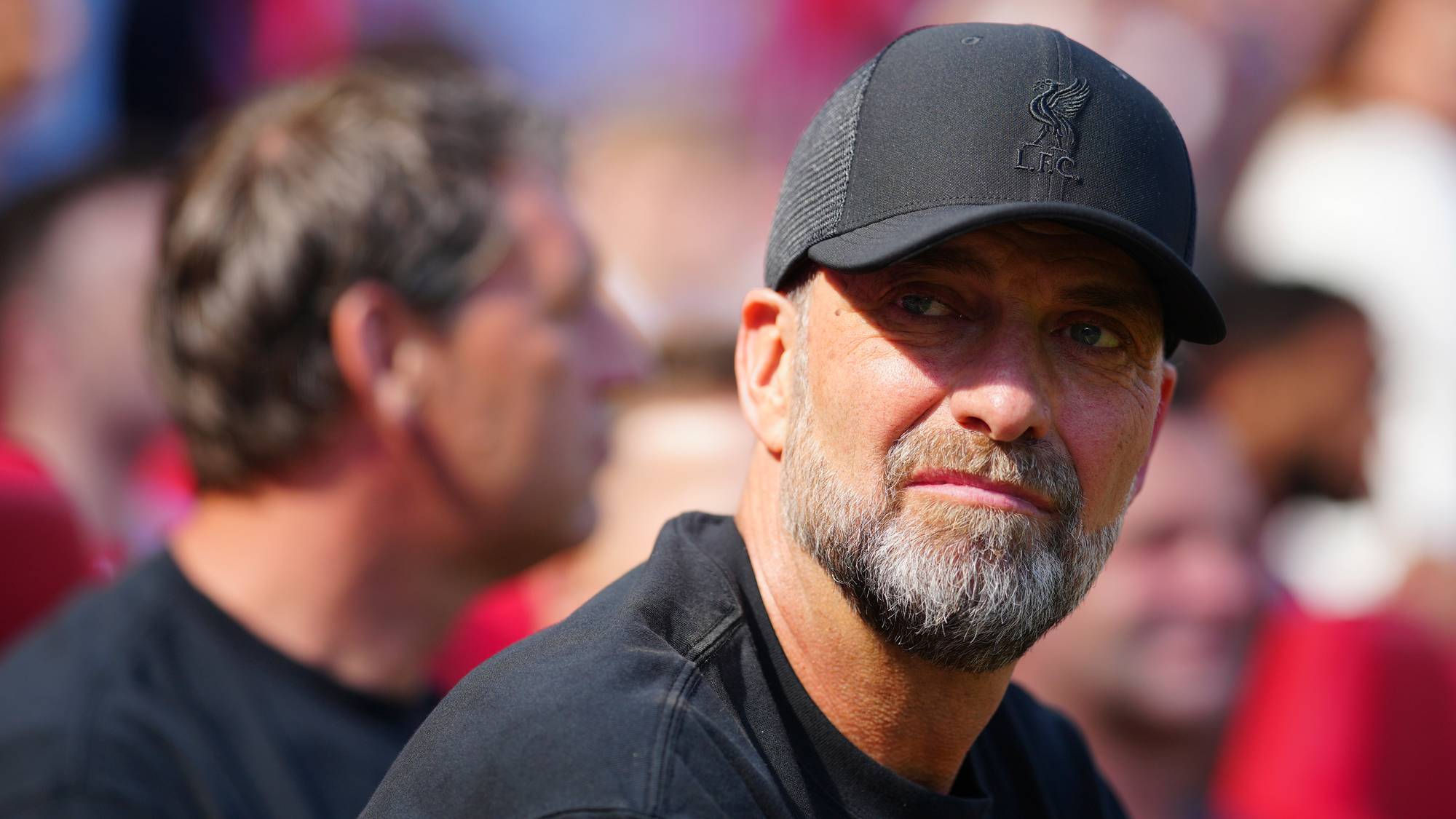 Jürgen Klopp med svart caps og t-skjorte ser mot siden mens han sitter på en fotballarena. (Bildebeskrivelsen er laget av en KI-tjeneste)