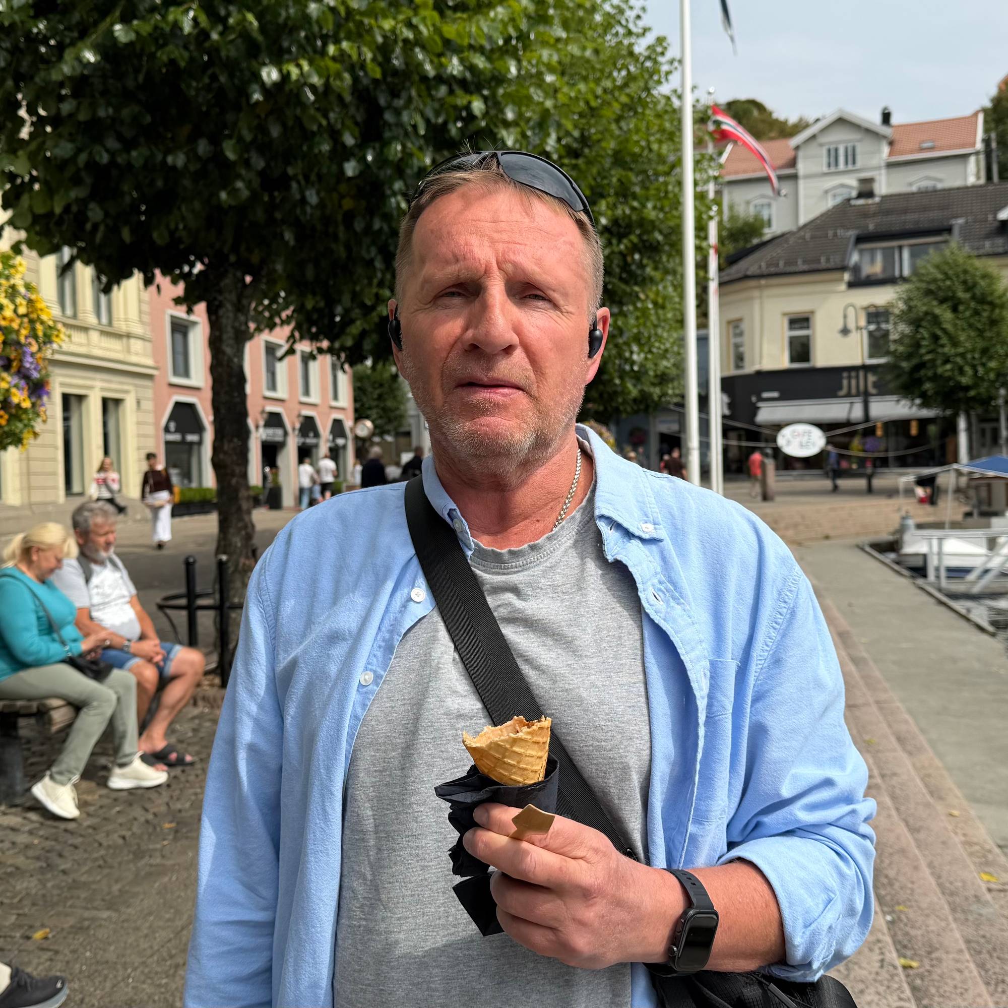 Mann i blå genser med iskrem, brygge og by i bakgrunnen. 