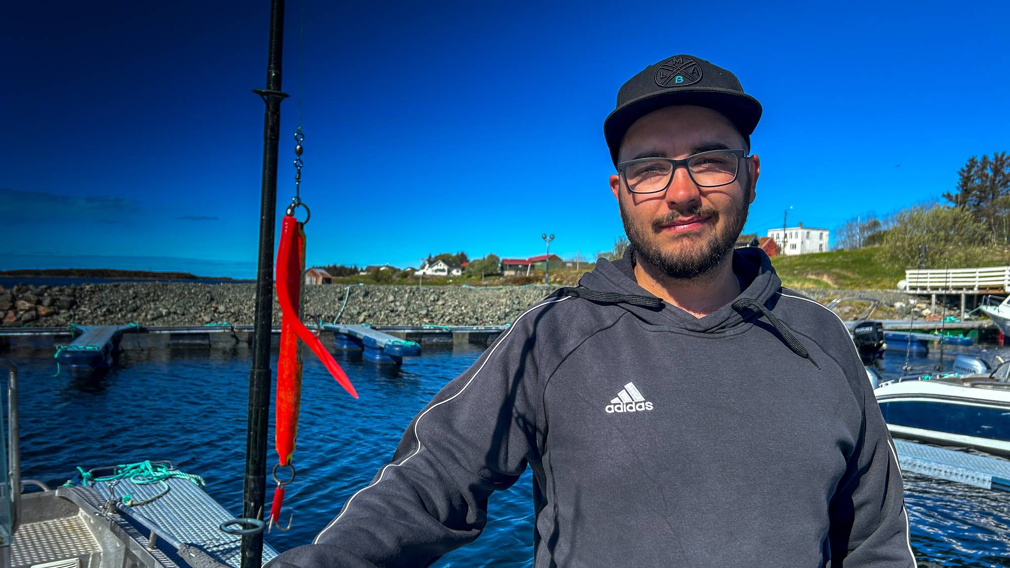 Mann med fiskeutstyr står på en båt. Han er iført en hettegenser og caps. Bak ham er det en småbåthavn med hus i bakgrunnen. En rød sluk henger fra fiskestangen. (Bildebeskrivelsen er laget av en KI-tjeneste)