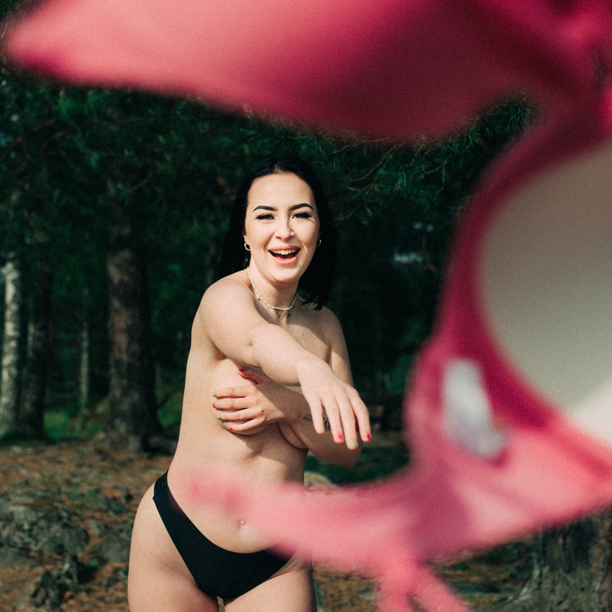 Maia (21) smiler stort mens hun kaster den rosa bikinitoppen mot kamera. Hun har venstre arm over brystene.