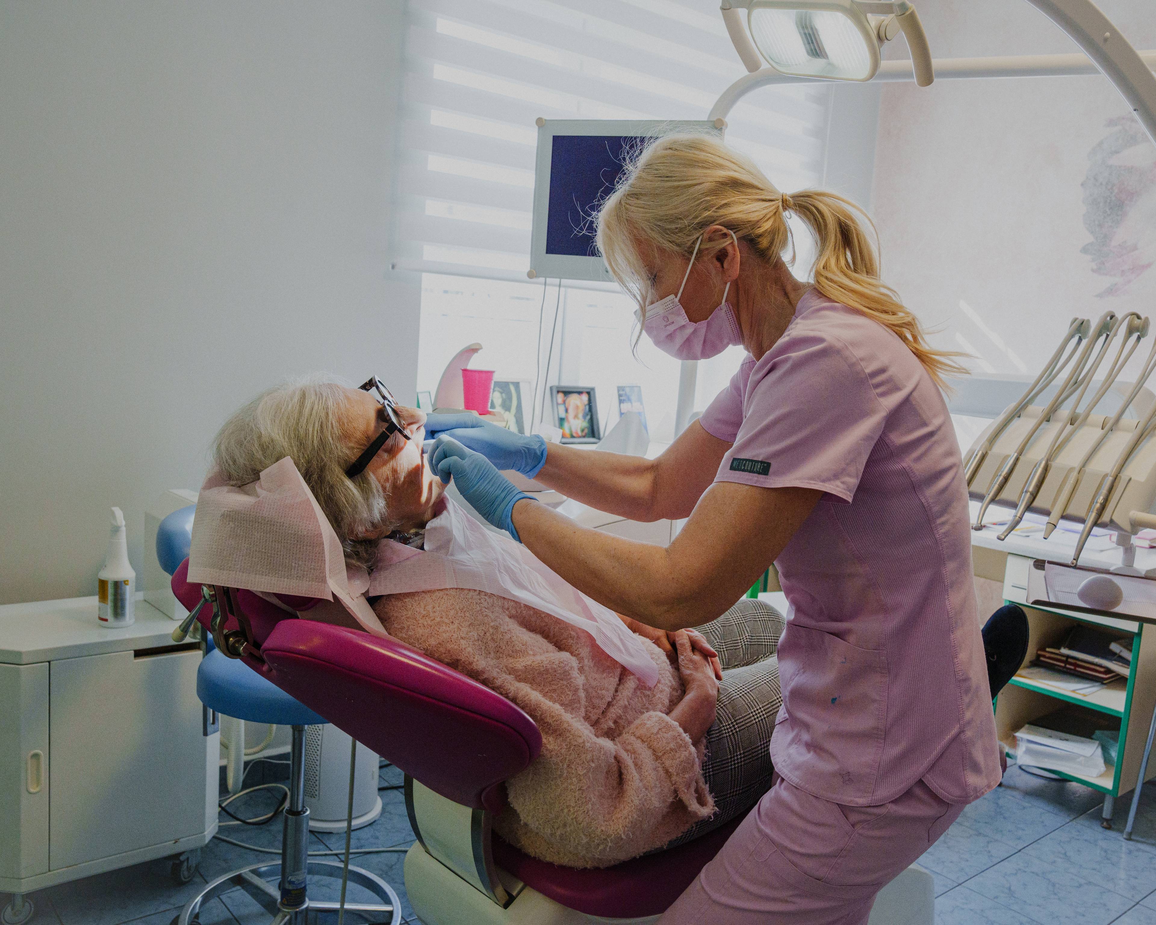 Magdalena ligger i en tannlegestol med solbriller på. En tannlege i rosa uniform og blondt hår inspiserer munnen hennes