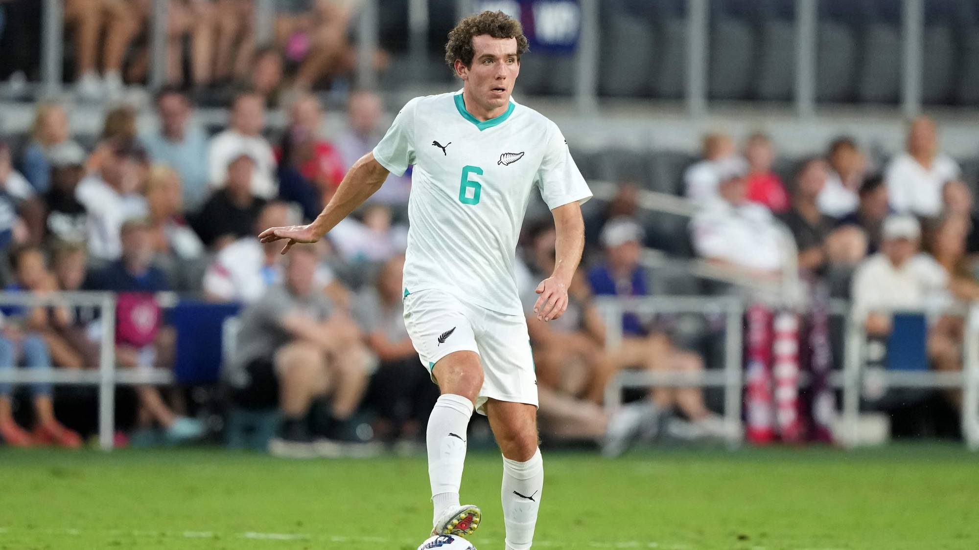 En fotballspiller i hvit drakt med nummer 6 dribler på gress. Spilleren har krøllete brunt hår. Tilskuere sitter på tribunen i bakgrunnen. (Bildebeskrivelsen er laget av en KI-tjeneste)