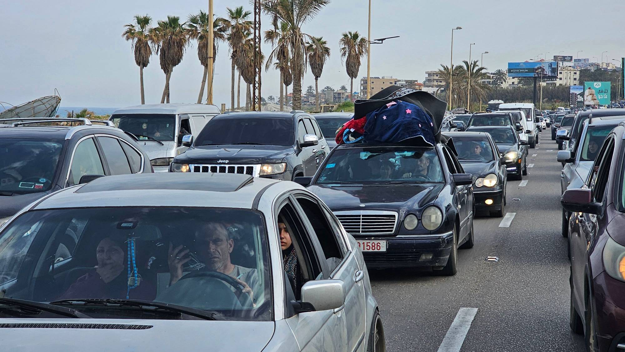 Lang kø av biler står fast langs en vei med palmetrær. En mann og en kvinne sitter i forsetet på en sølvfarget bil. Bagasje er stablet på taket av en svart Mercedes. En annen svart bil ligger i veien. (Bildebeskrivelsen er laget av en KI-tjeneste)