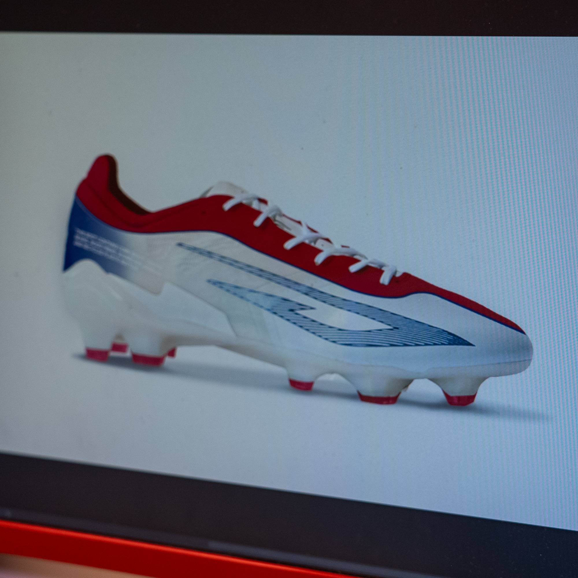 En dataskjerm viser et bilde av en fotballsko. Bildet er en del av et designprogram på en datamaskin. Skoen er hvit med røde og blå detaljer. Skjermbildet viser også menyer og verktøy fra designprogramvaren. (Bildebeskrivelsen er laget av en KI-tjeneste)